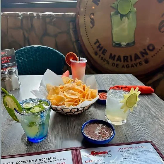 Table with two cocktails, a basket of tortilla chips, salsa, and a pink drink. In the background, a large barrel with a logo for 'The Mariano' tequila brand.