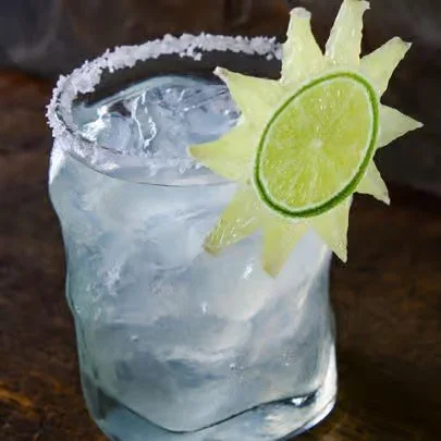 A glass of clear cocktail garnished with a slice of lime and a pineapple-shaped fruit decoration, with a salted rim and ice