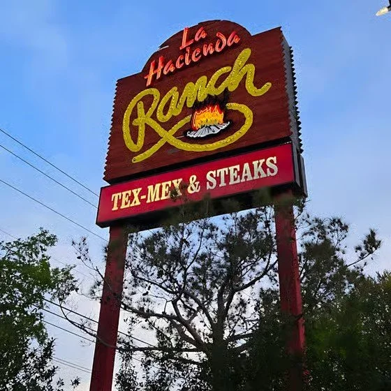 Sign for La Hacienda Ranch restaurant featuring Tex-Mex cuisine and steaks, with the restaurant's name in large yellow and red letters, and a depiction of a fire and steak.