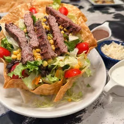 A taco salad with lettuce, cherry tomatoes, sliced steak, corn, and crispy tortilla bowl, served with sides of sour cream and shredded cheese.