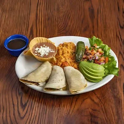 Plate with four rolled tortillas, rice, refried beans with cheese, avocado slices, lime, cucumber, chopped vegetables, and a small bowl of salsa on a wooden table.