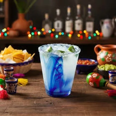 A glass of blue and clear cocktail with lime slices on a rustic wooden table with Mexican decorations, chips, and snacks in the background.