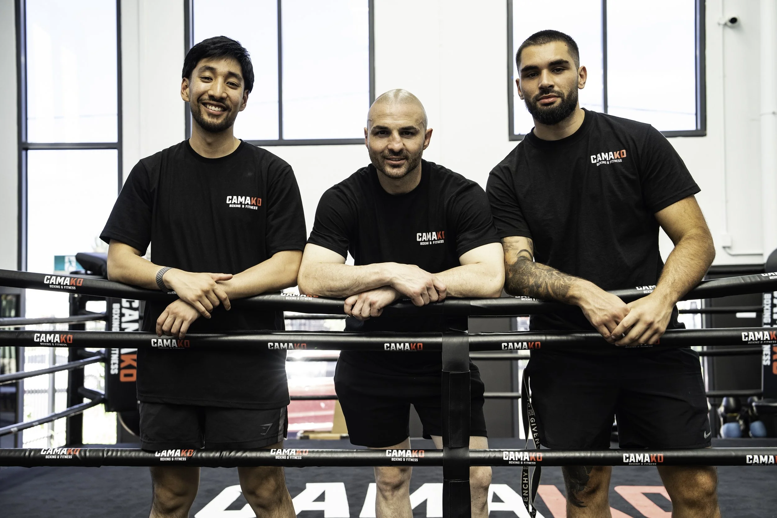 Three men in black t-shirts with 'CAMAKO Boxing & Fitness' logo standing inside a boxing gym, leaning on a boxing ring.