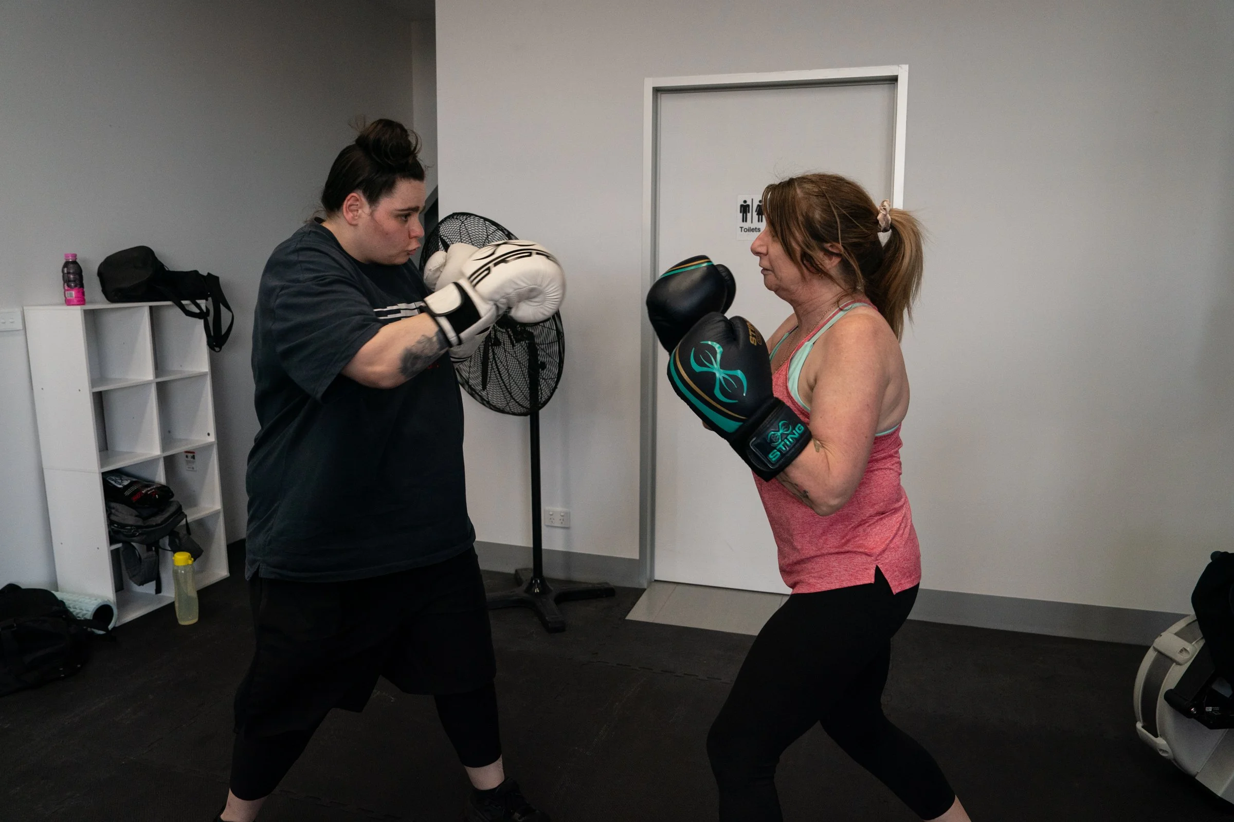 Two women practicing boxing indoors, both wearing boxing gloves and standing in fighting stances facing each other.