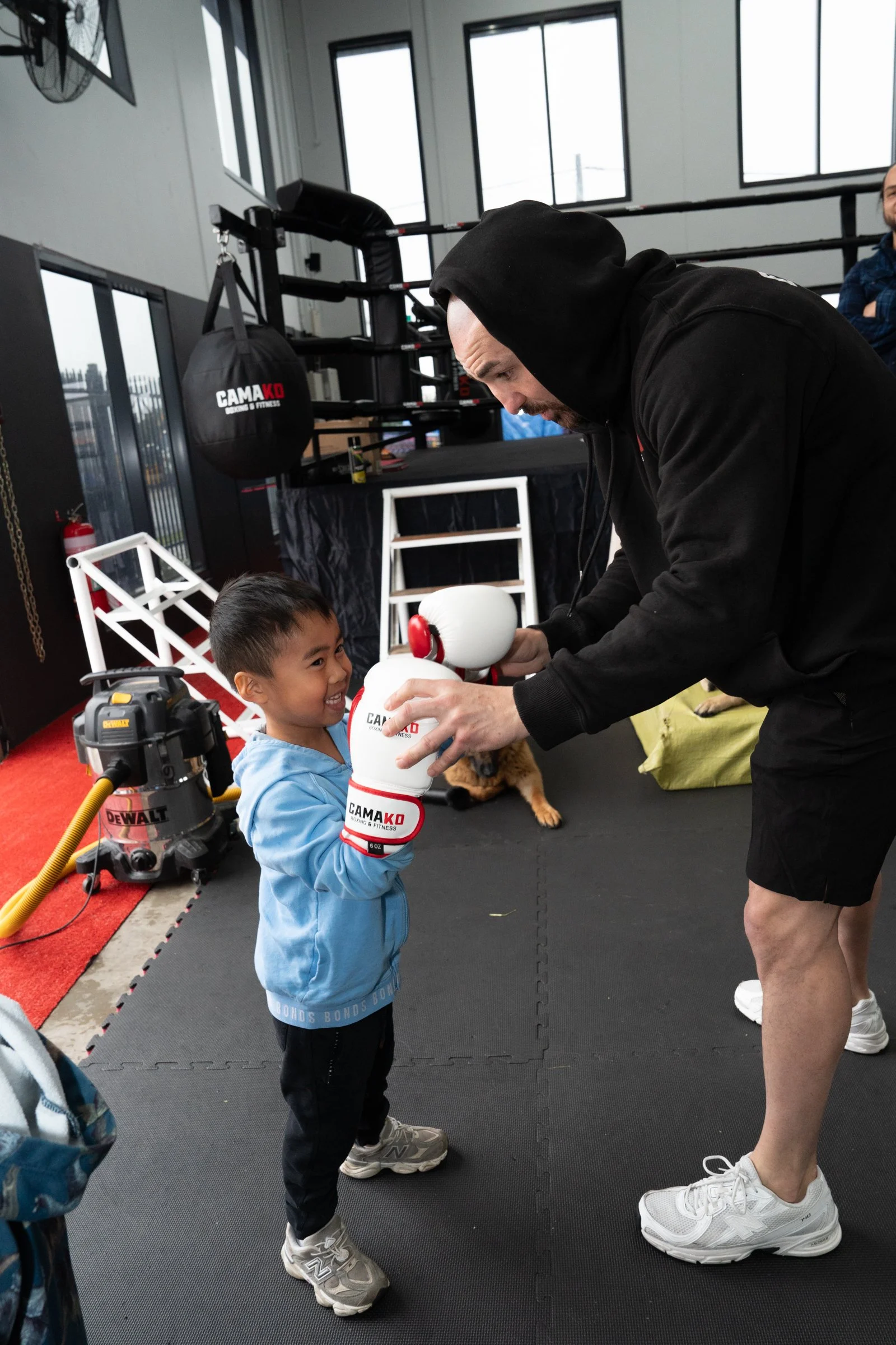 A man in a black hoodie and shorts teaching boxing to a young boy in a gym. The boy is smiling and wearing boxing gloves, while the man is holding a focus mitt. Other gym equipment and a dog are visible in the background.