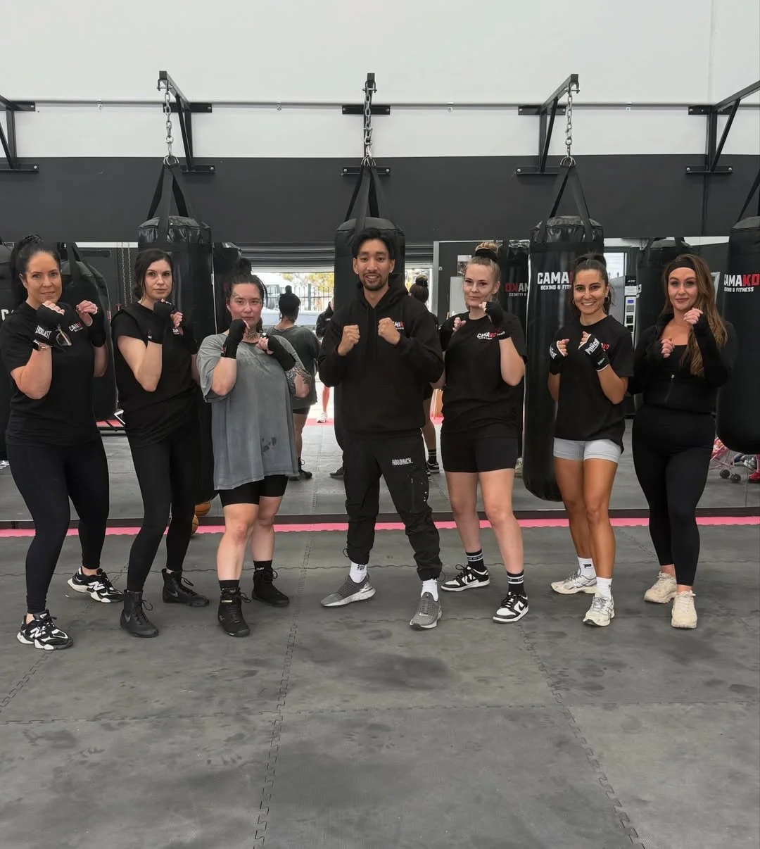 Group of seven women and one man in gym wearing boxing gloves, standing in front of punching bags, smiling at camera.