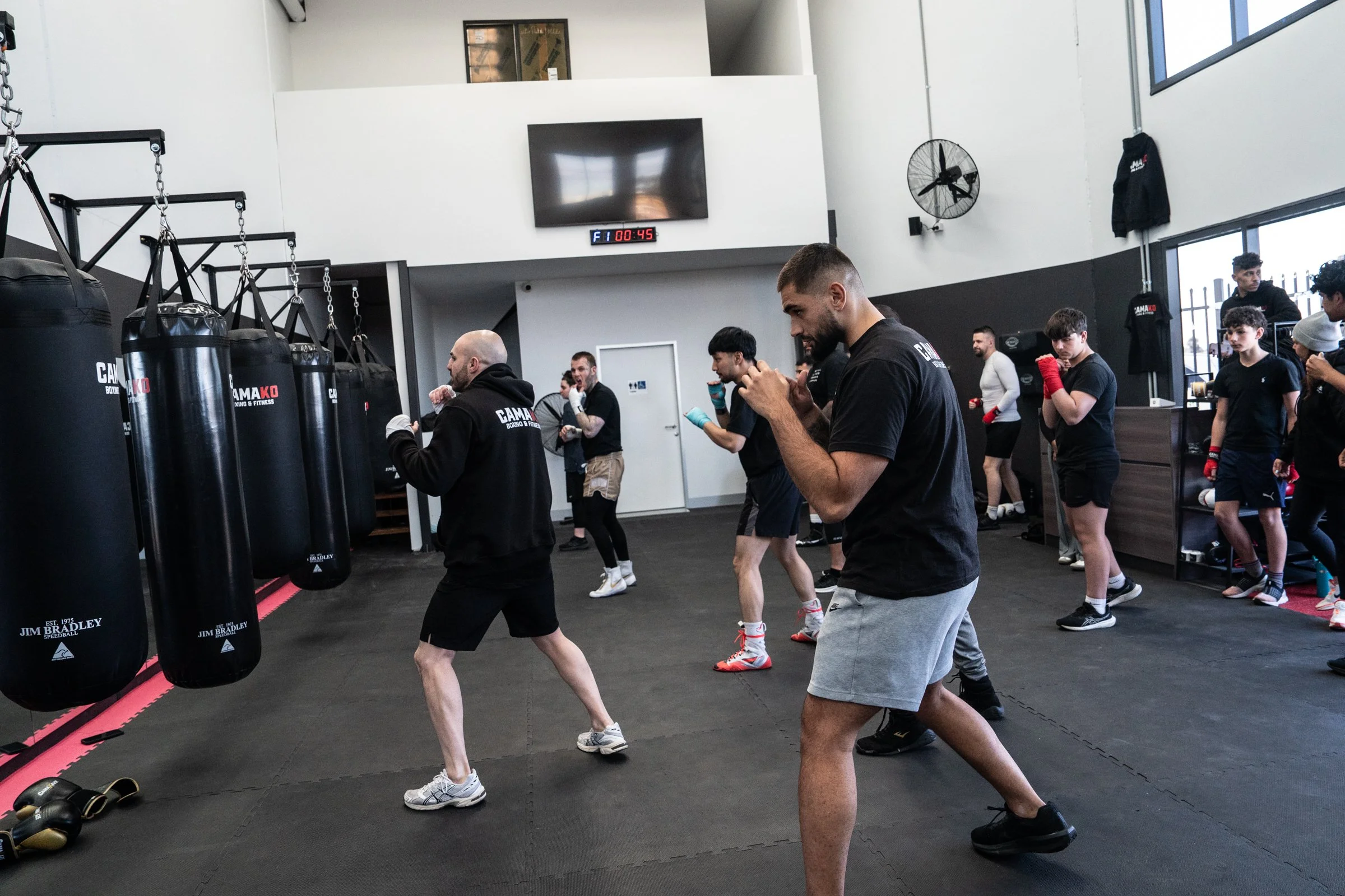 People practicing boxing in a gym, facing punching bags, with some role models in the background and a large television screen showing a timer.