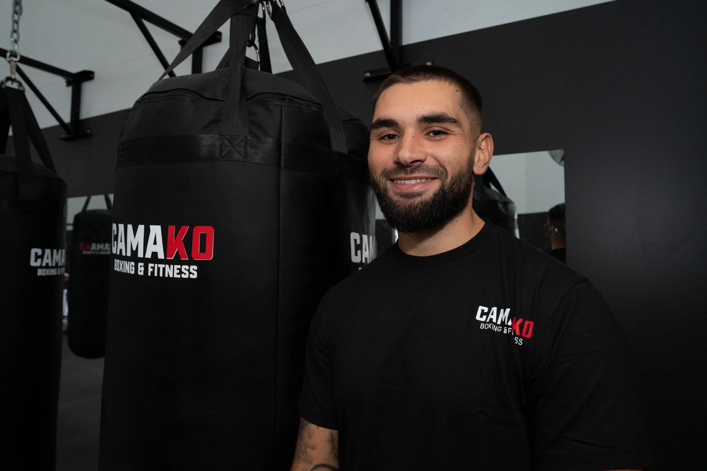 A man with a beard smiling at the camera, standing in a boxing gym near hanging black punching bags with 'CAMA KID Boxing & Fitness' logos.