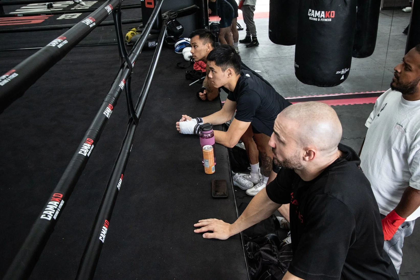 A group of boxers and trainers sit and kneel on the floor of a boxing gym, resting during a training session. Some are leaning forward, and boxing gloves and equipment are visible in the background.