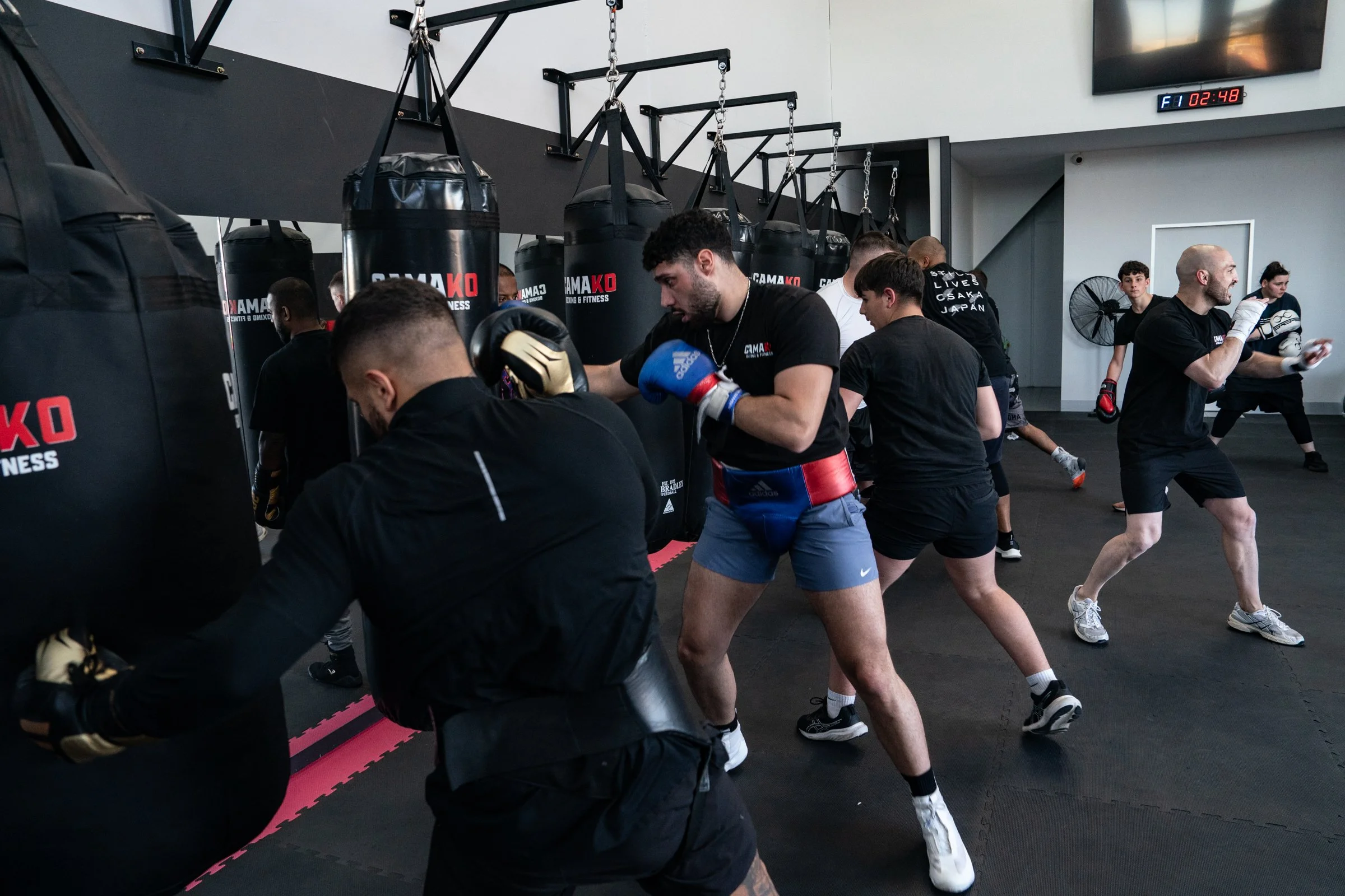 A group of people practicing boxing in a gym, wearing gloves and athletic clothing, with punching bags and a digital clock on the wall.