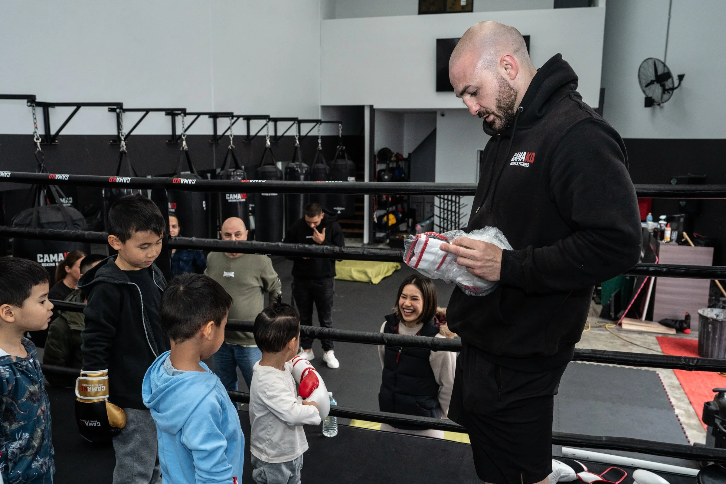 A group of children and adults gather around a man in a boxing gym. The man, wearing a black hoodie, is handing a package to a young girl wearing boxing gloves. Punching bags are visible in the background.