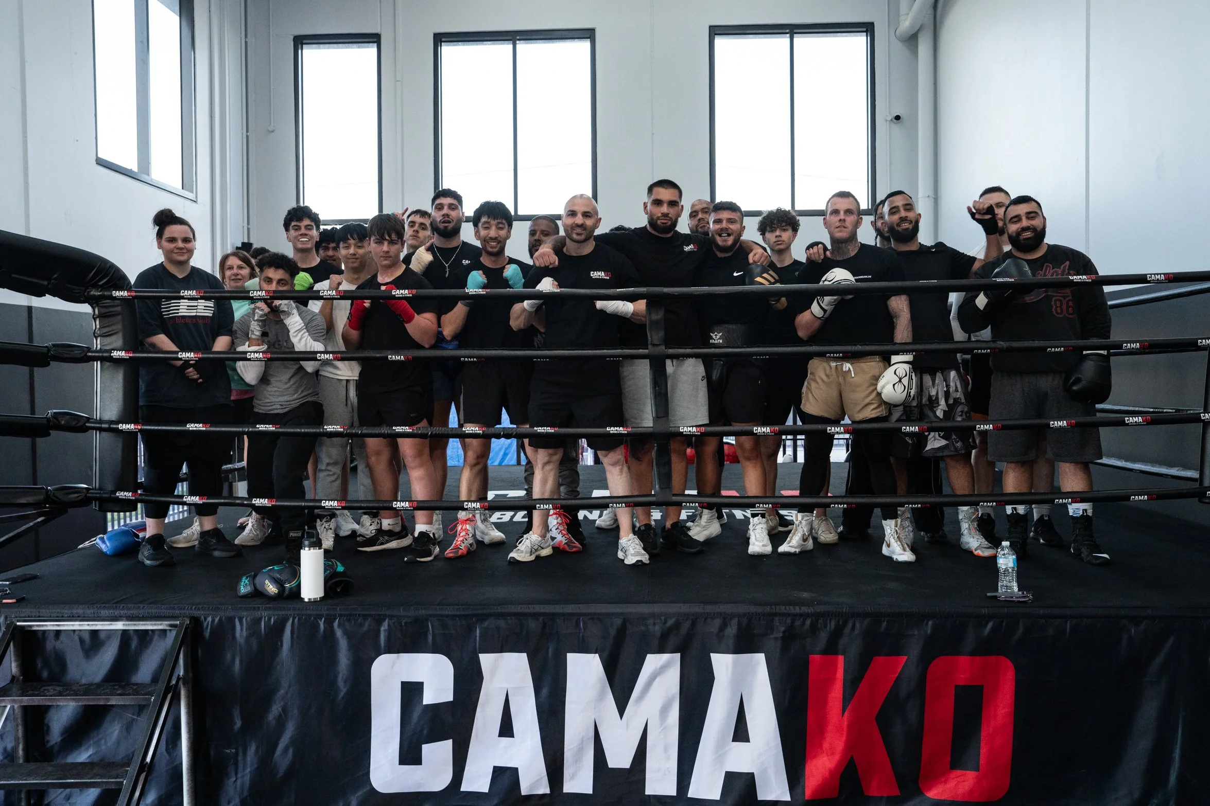 Group of diverse athletes in boxing gym, standing behind boxing ring, some wearing boxing gloves, posing for photo, training equipment on floor.
