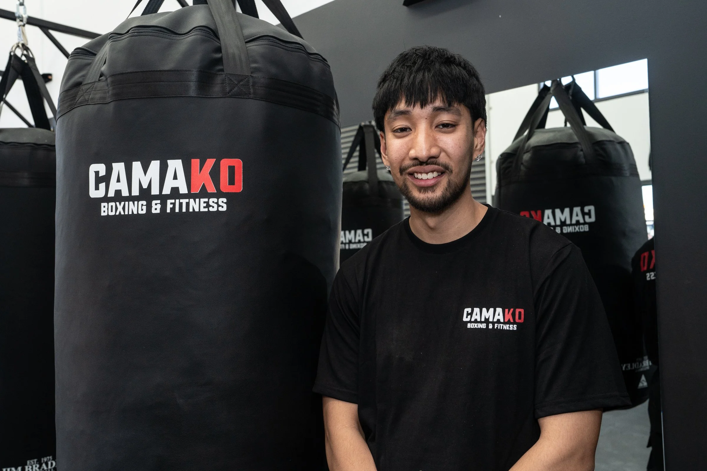 A young man with black hair and a short beard near a punching bag in a gym. He is smiling and wearing a black t-shirt with the logo 'CAMA KO Boxing & Fitness.' Multiple punching bags are visible in the background.
