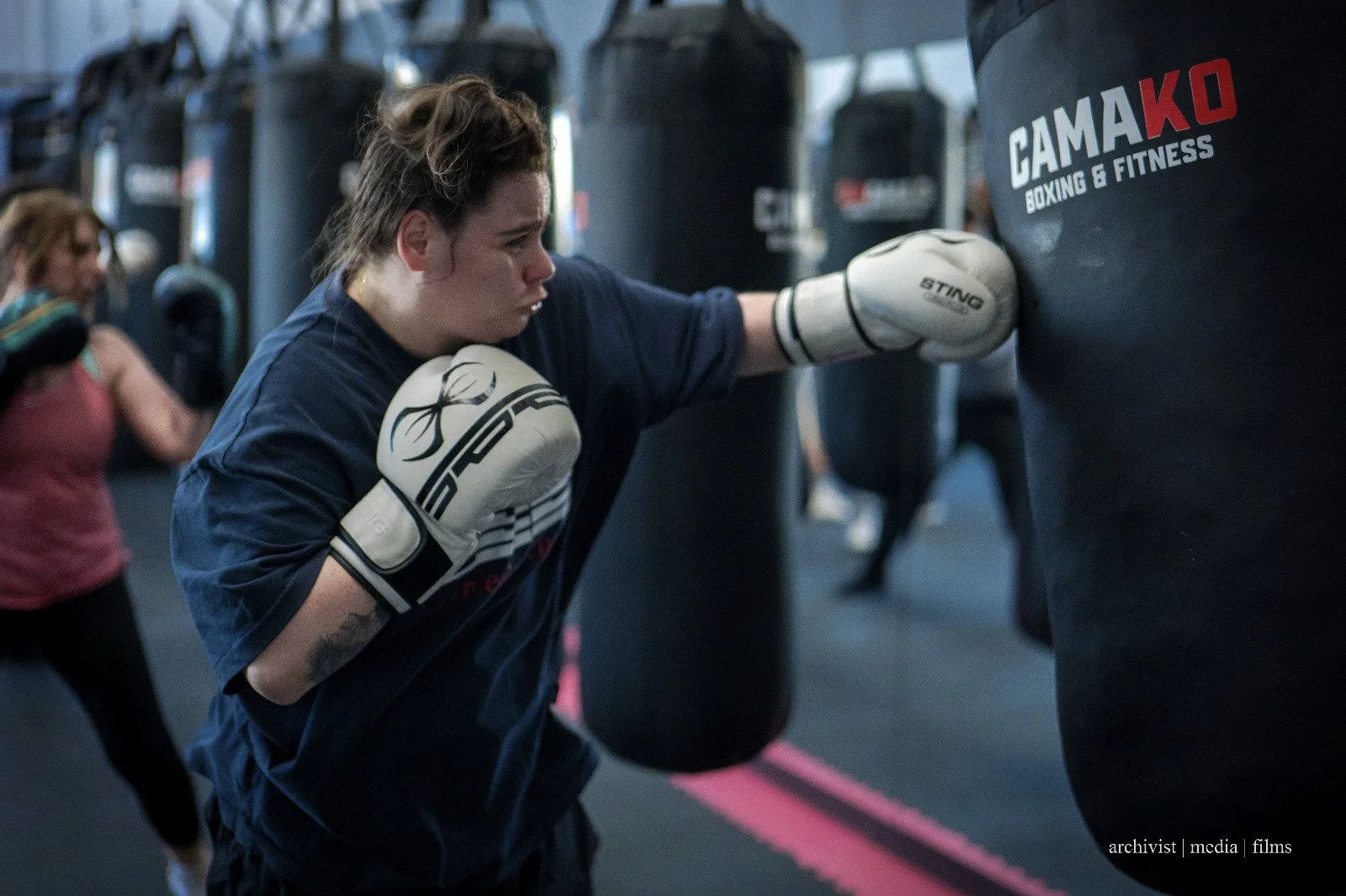 A woman with tattoos wearing boxing gloves and a blue shirt trains by punching a black punching bag with the word 'GAMAKO' on it at a boxing gym.