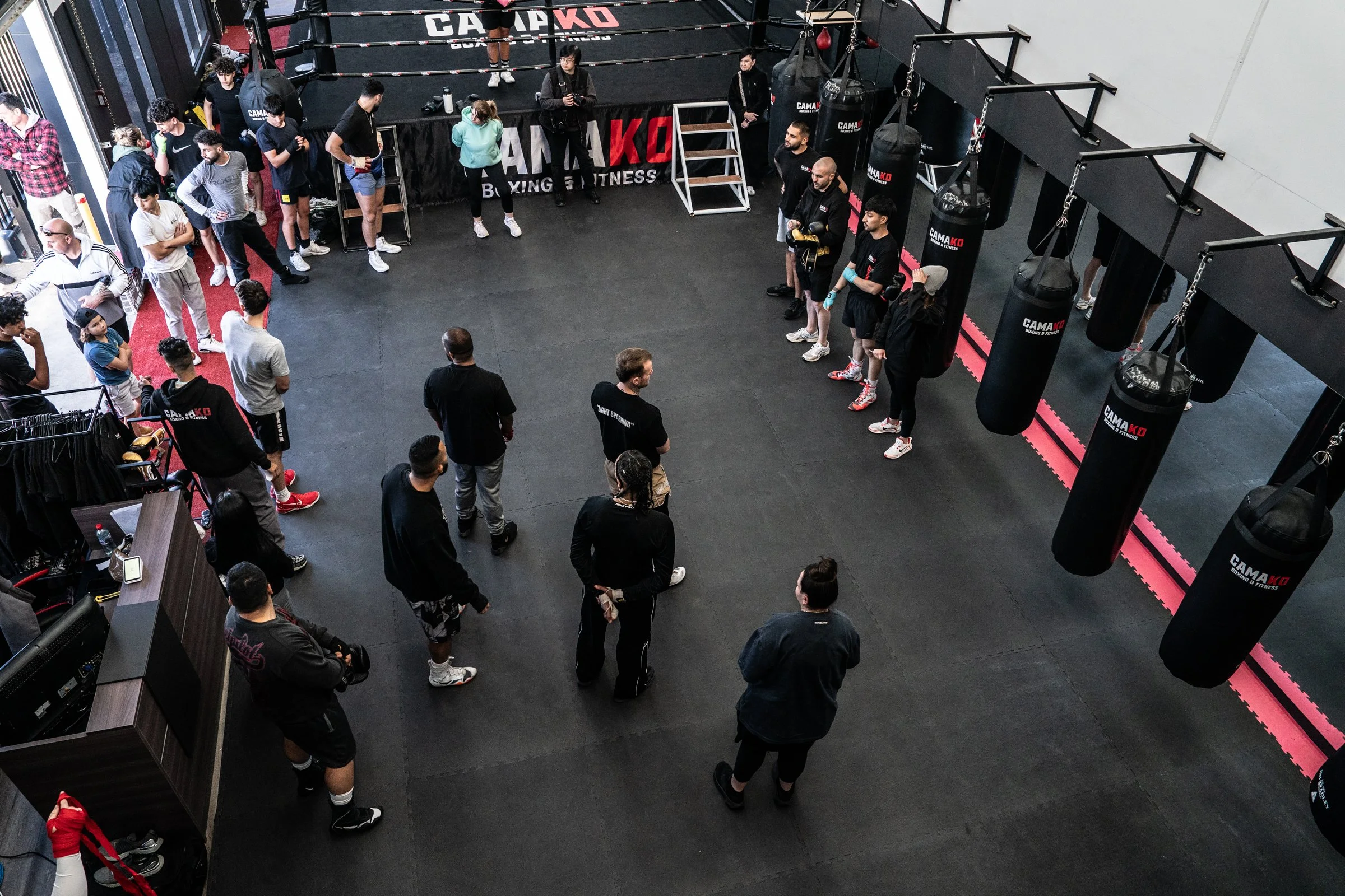 People gathered inside a boxing gym with punching bags, a boxing ring, and trainers, possibly waiting for training or a class.