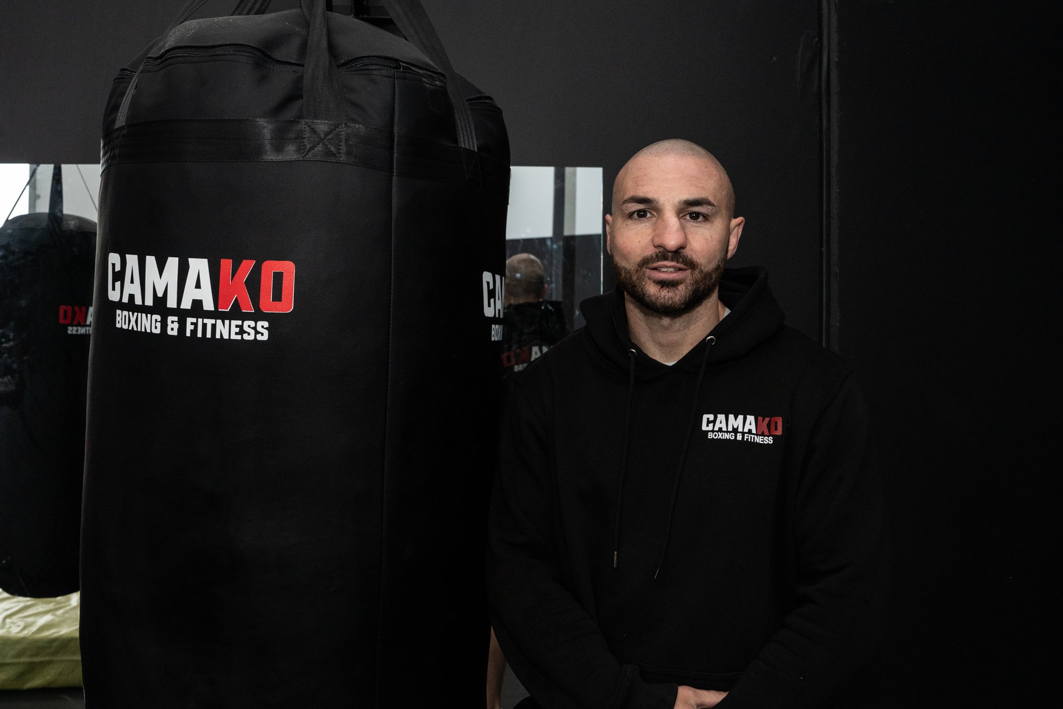 A man with a bald head and beard wearing a black hoodie with a logo that says 'CAMA K.O Boxing & Fitness' stands next to a punching bag inside a gym.