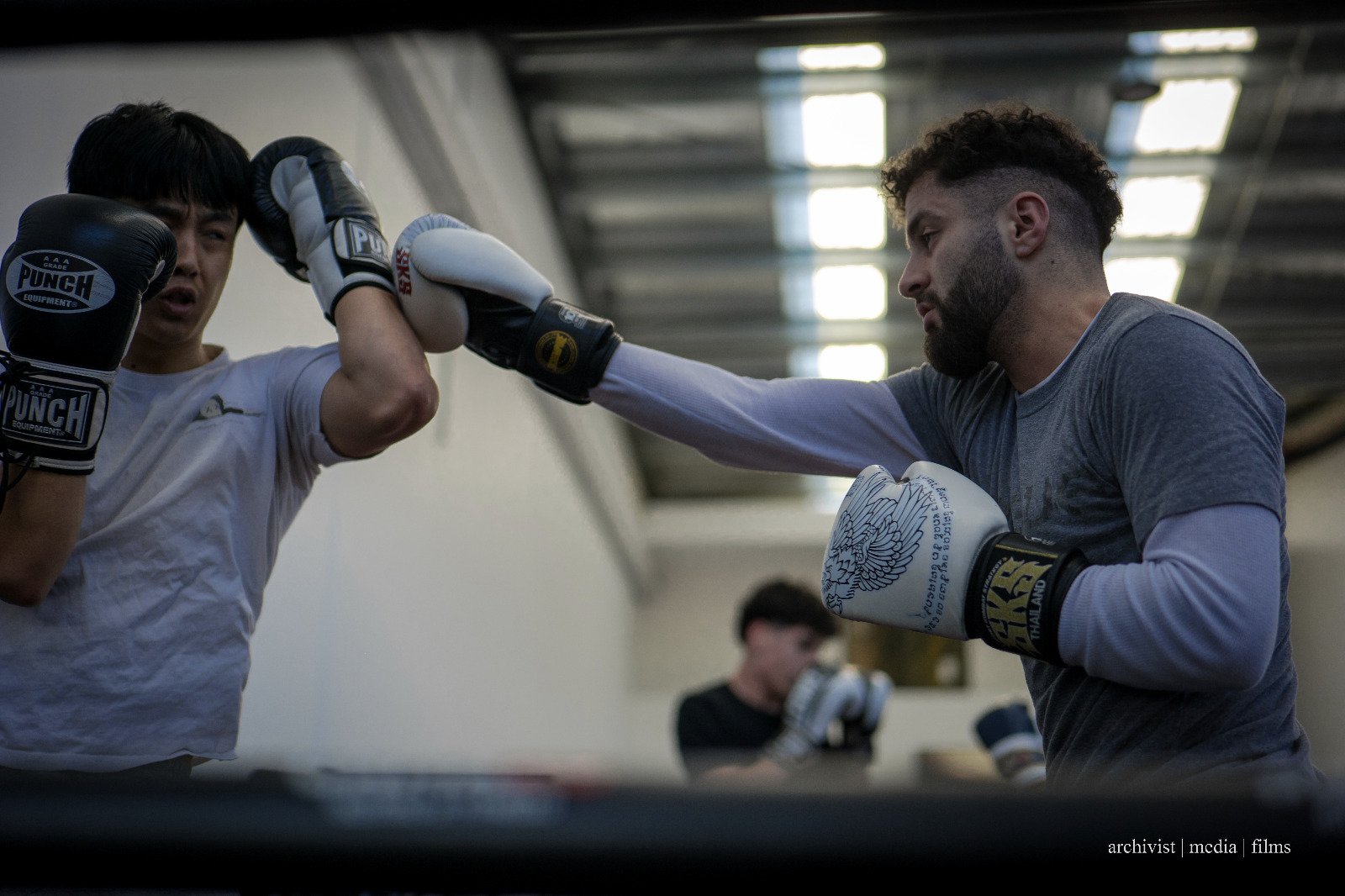 Two men sparring in a boxing gym, with one throwing a punch toward the other's face while wearing boxing gloves and athletic clothing.