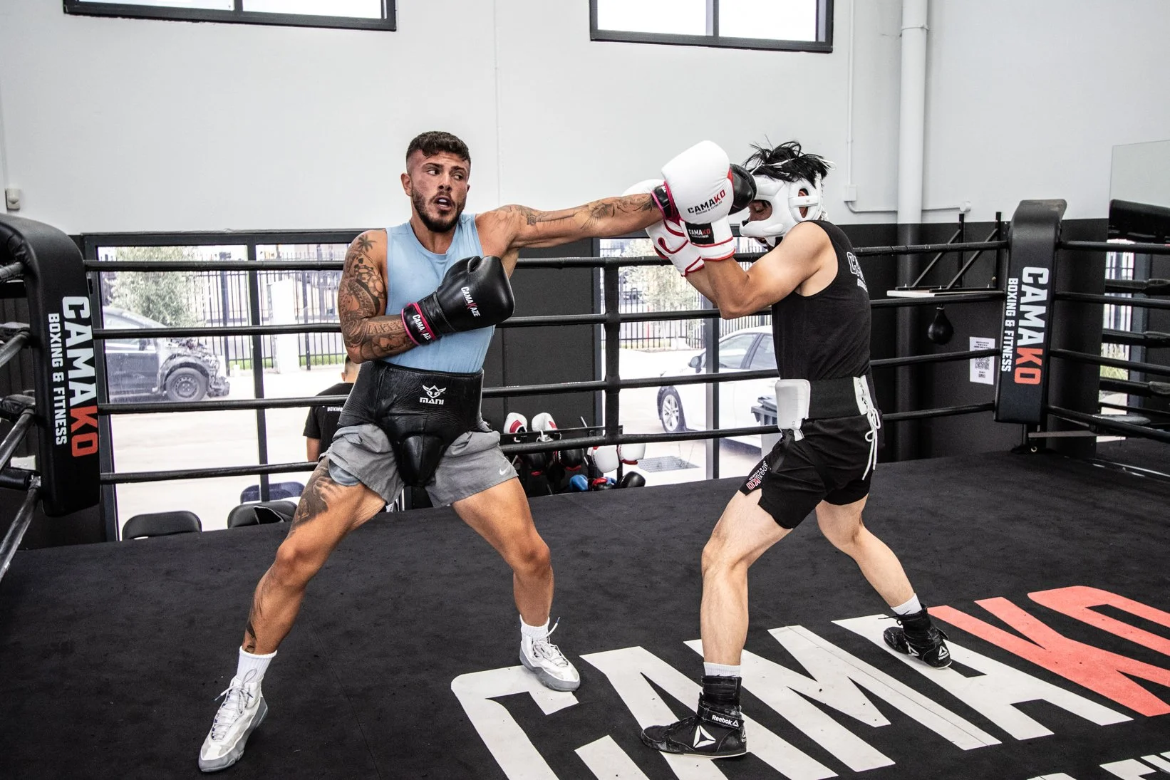 Two men boxing in a gym. One man is throwing a punch towards the other, who is blocking with his gloves. They are wearing boxing gloves, shorts, and protective gear. The gym has a boxing ring with canvas flooring and padded ropes.