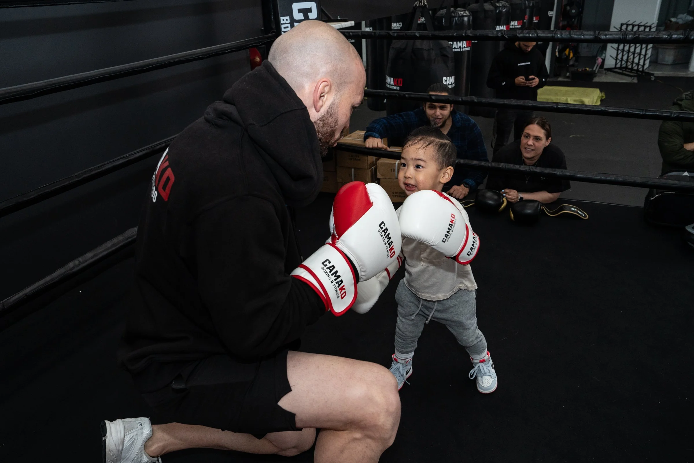A muscular man with a shaved head and beard, wearing black shorts and white sneakers, kneeling in a boxing ring and holding a young boy wearing boxing gloves, grey sweatpants, and sneakers, while the boy looks up at the man. In the background, there are spectators and boxing equipment.