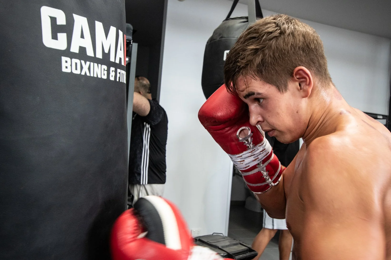 A shirtless young male boxer with red gloves practicing punches on a black punch bag in a gym, with a focused expression.