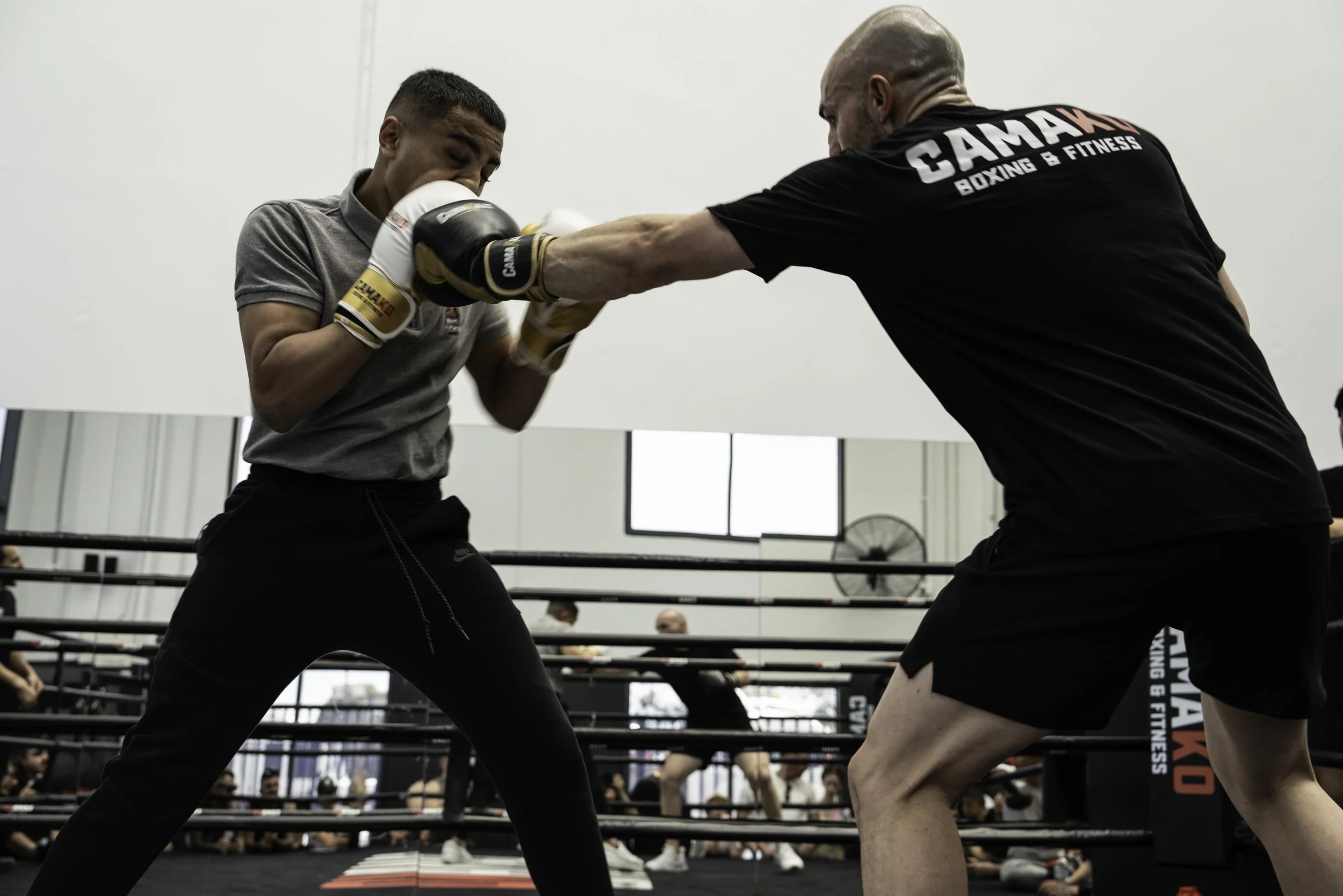 Two men sparring in a boxing ring, one wearing black shorts and a black shirt with 'GAMAX Boxing & Fitness' on the back, and the other in gray shirt and black pants. The man in the black shirt throws a punch while the other blocks or absorbs it with his gloves, with spectators watching behind them.