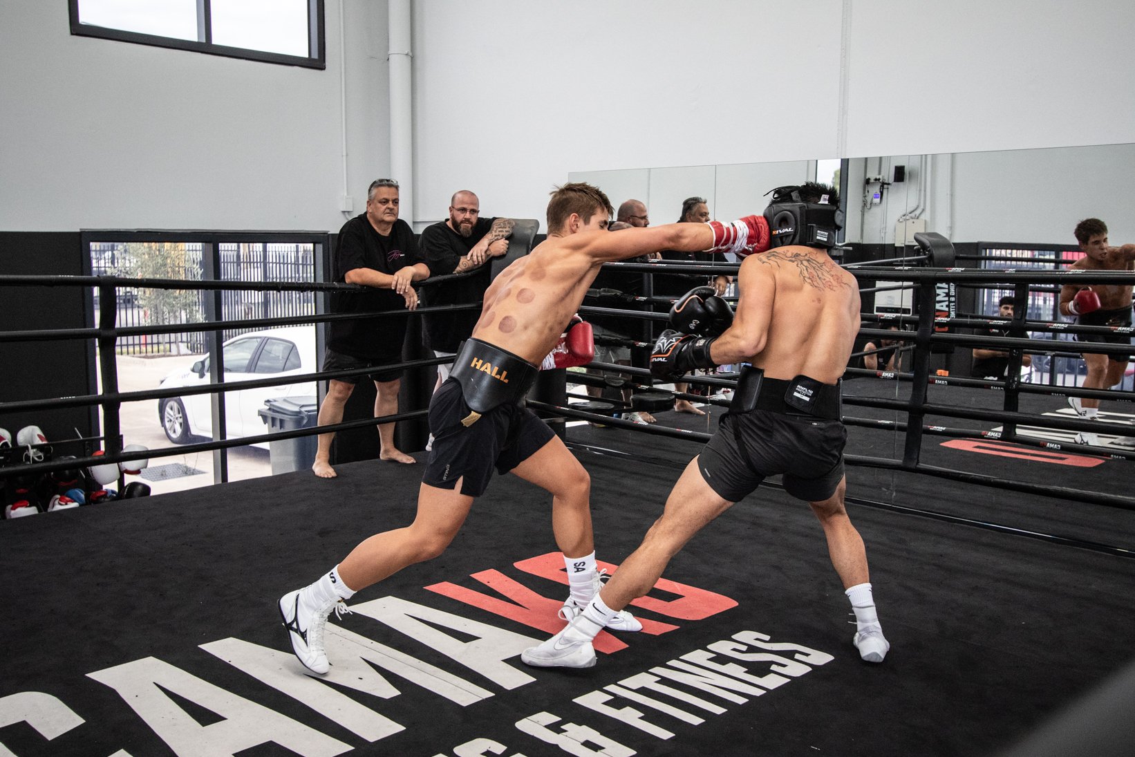 Two shirtless male fighters with boxing gloves engaged in a boxing match inside a ring, with five men observing from outside the ring. One fighter is throwing a punch while the other is blocking.