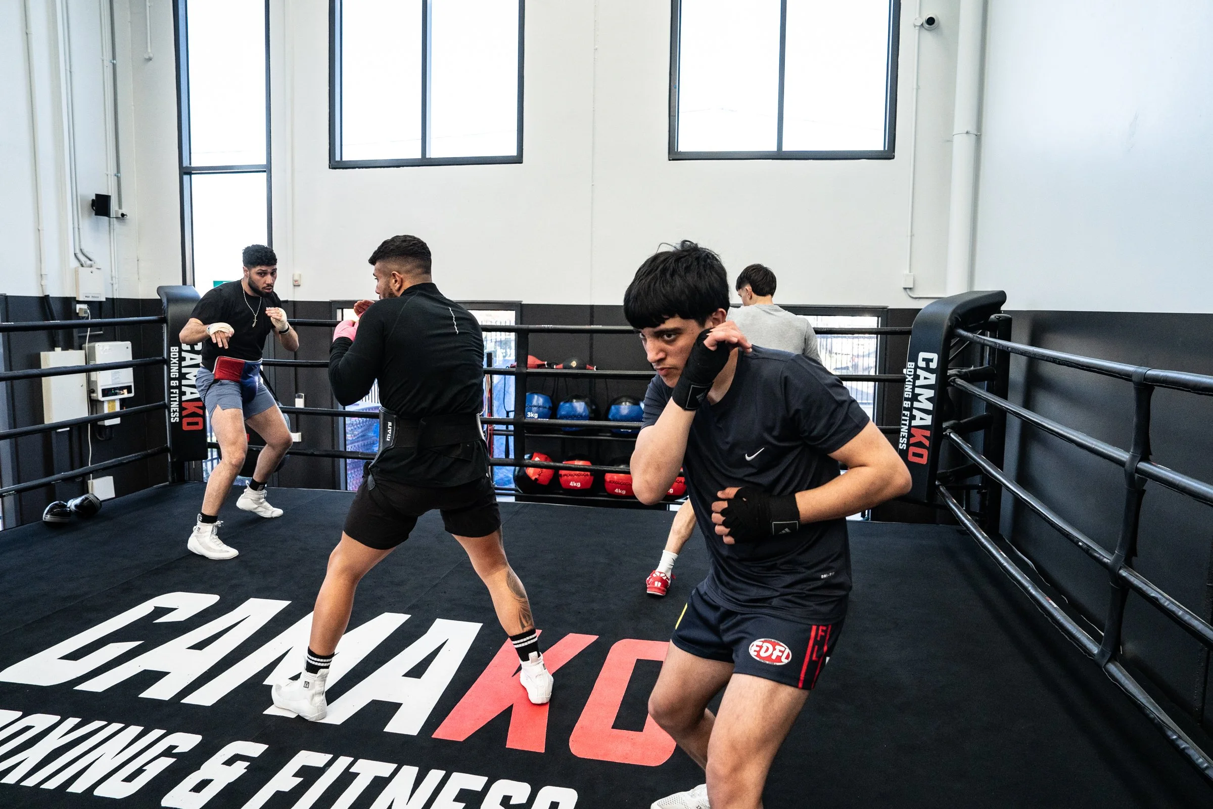 Two young men practicing boxing in a boxing ring, with a coach and another person in the background, inside a gym with large windows.