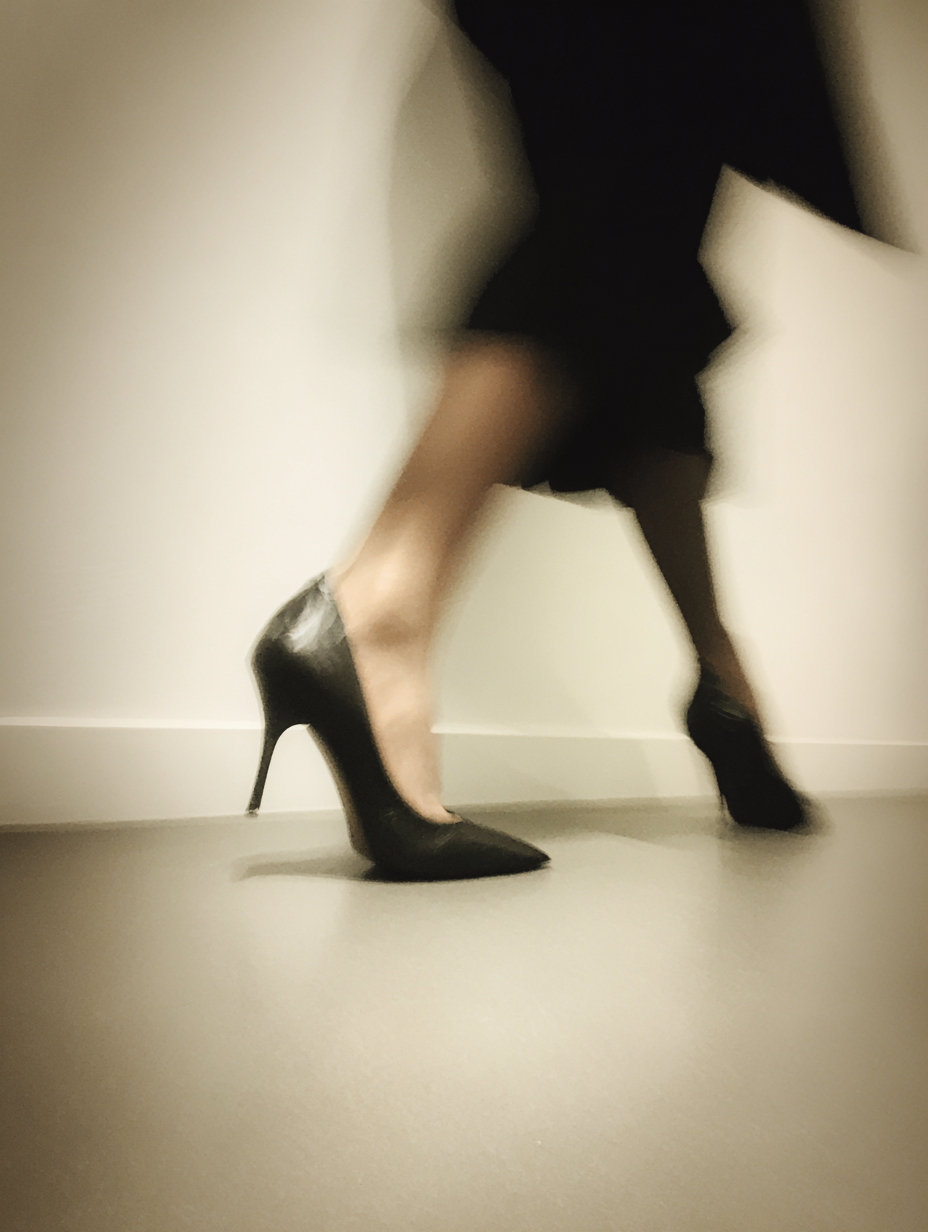Blurred photo of a person walking or dancing, wearing black high-heeled shoes and black clothing, against a light-colored wall and floor.