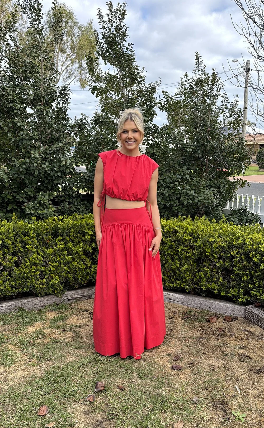 A woman wearing a bright red, two-piece outfit, standing outdoors in front of green bushes and trees, with a cloudy sky and suburban houses in the background.