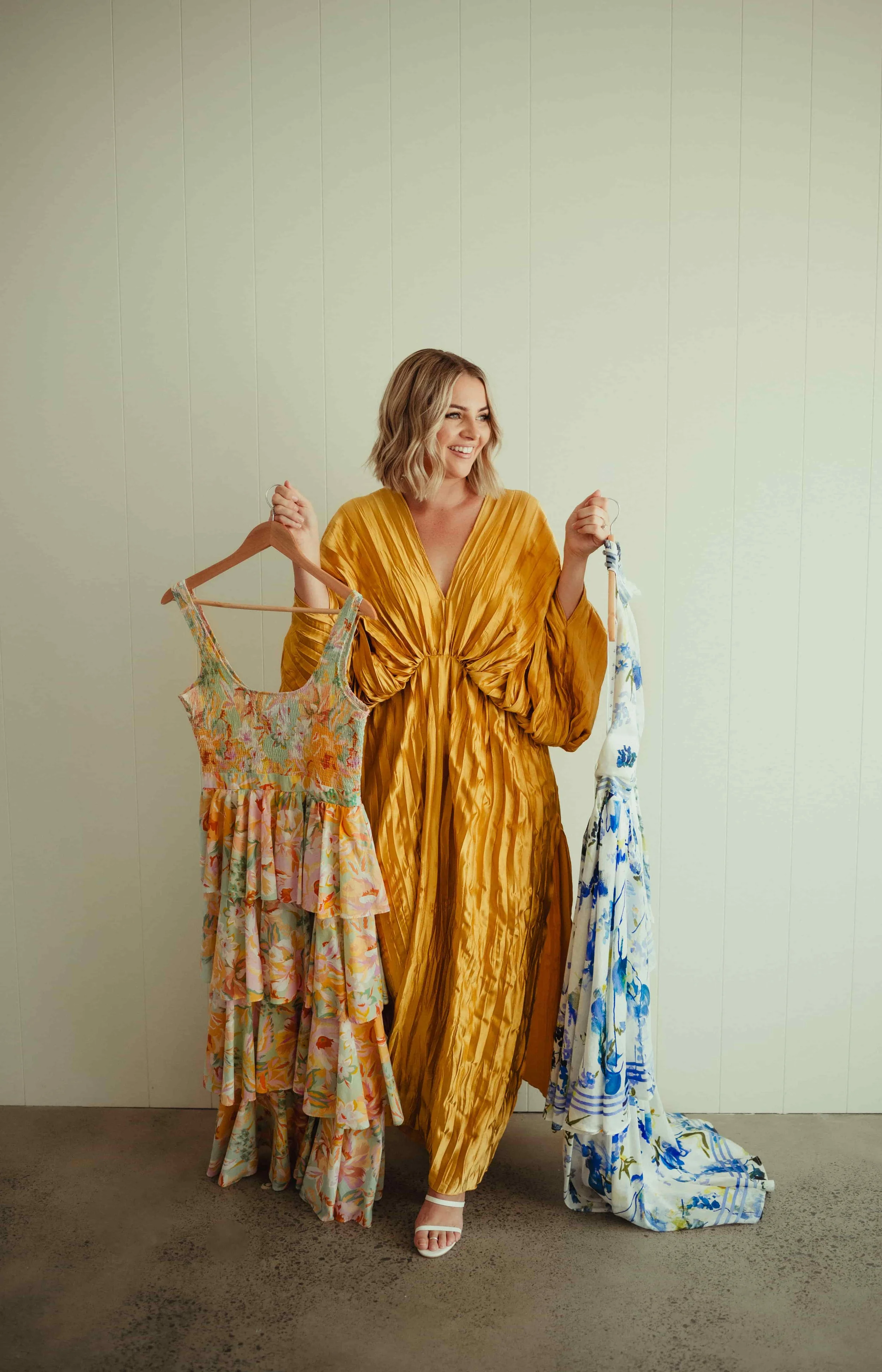 Woman in a gold dress holding colorful dresses on hangers, standing against a plain wall.