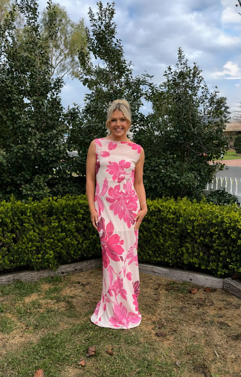 A woman in a sleeveless, long white dress with pink floral patterns stands outdoors in front of green bushes and trees, smiling at the camera with a cloudy sky overhead.