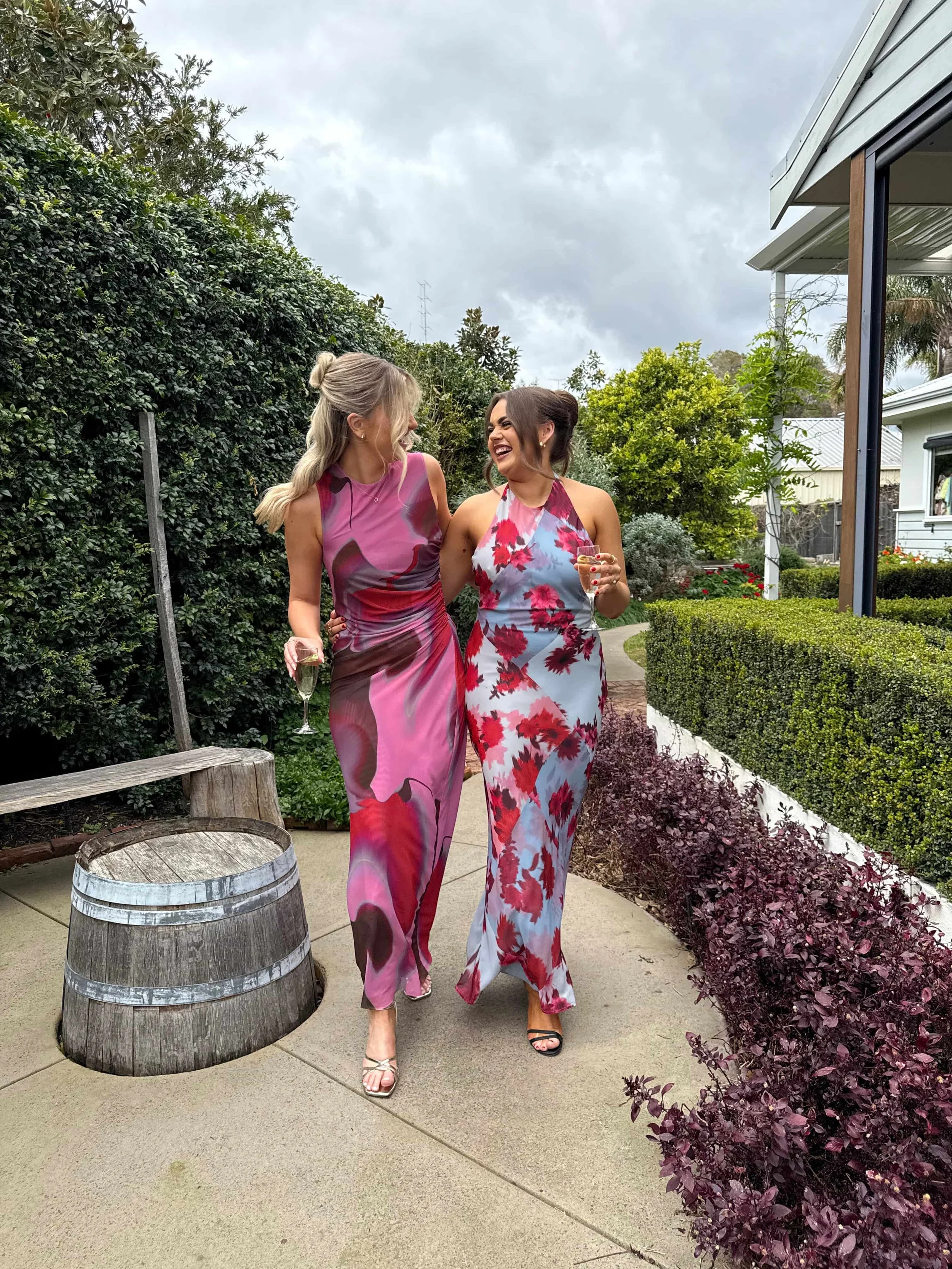 Two women walking outdoors, laughing and holding glasses of champagne, dressed in colorful floral dresses.