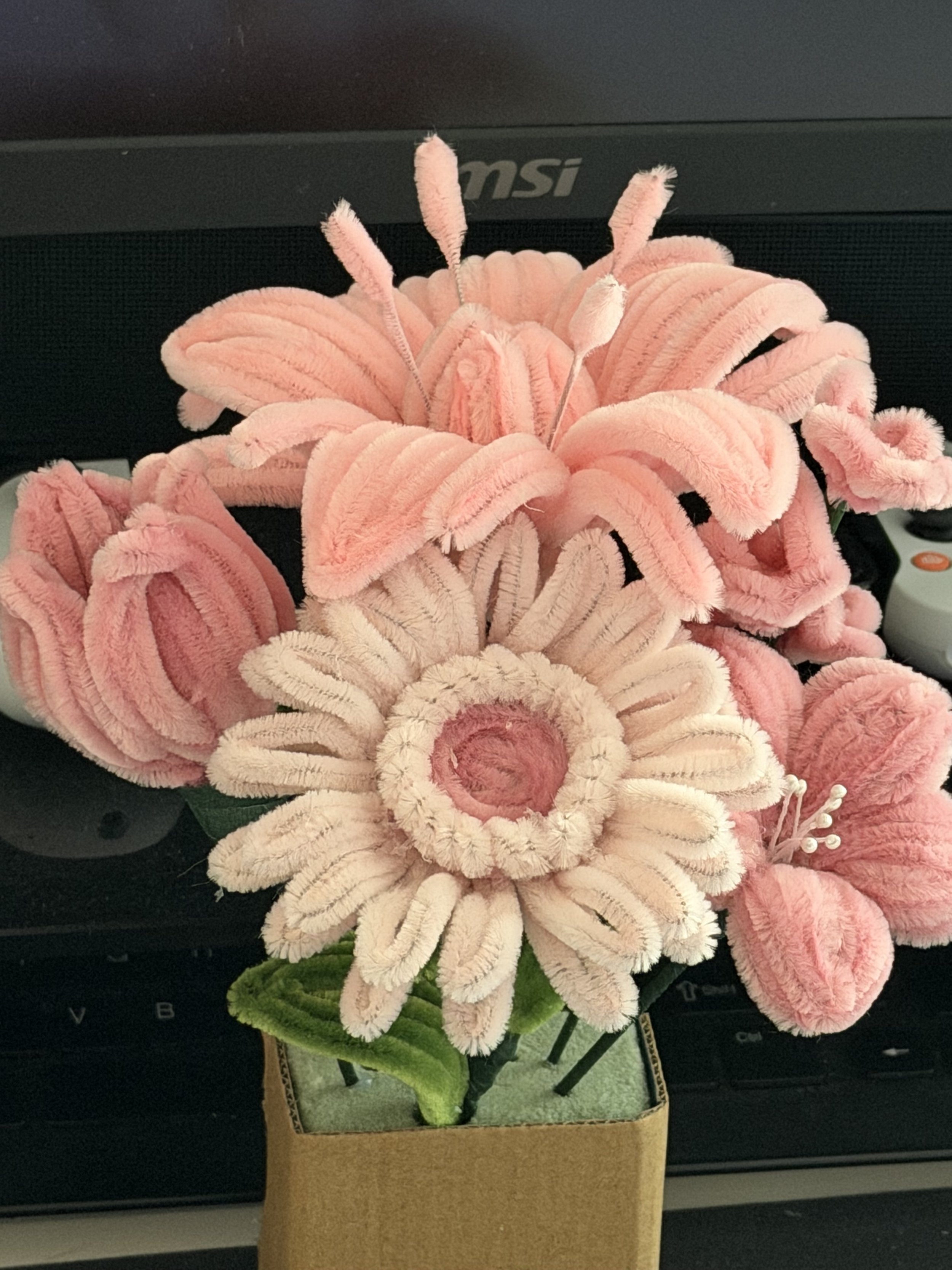 A pink, plush artificial flower arrangement in a small, square, beige pot on a black surface, with a gray computer keyboard and monitor in the background.