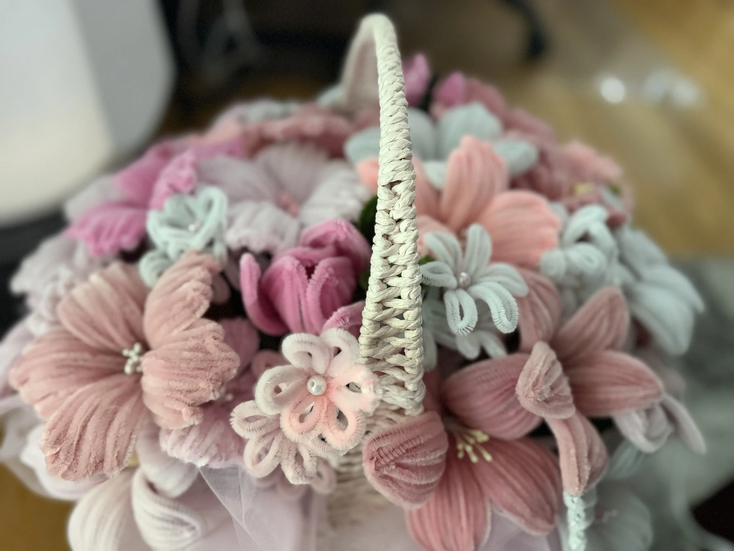 Close-up of a decorative arrangement made of pastel-colored fabric or yarn, with a focus on a central braided handle or stem, surrounded by curled and embroidered fabric flowers in pink, white, and light blue.