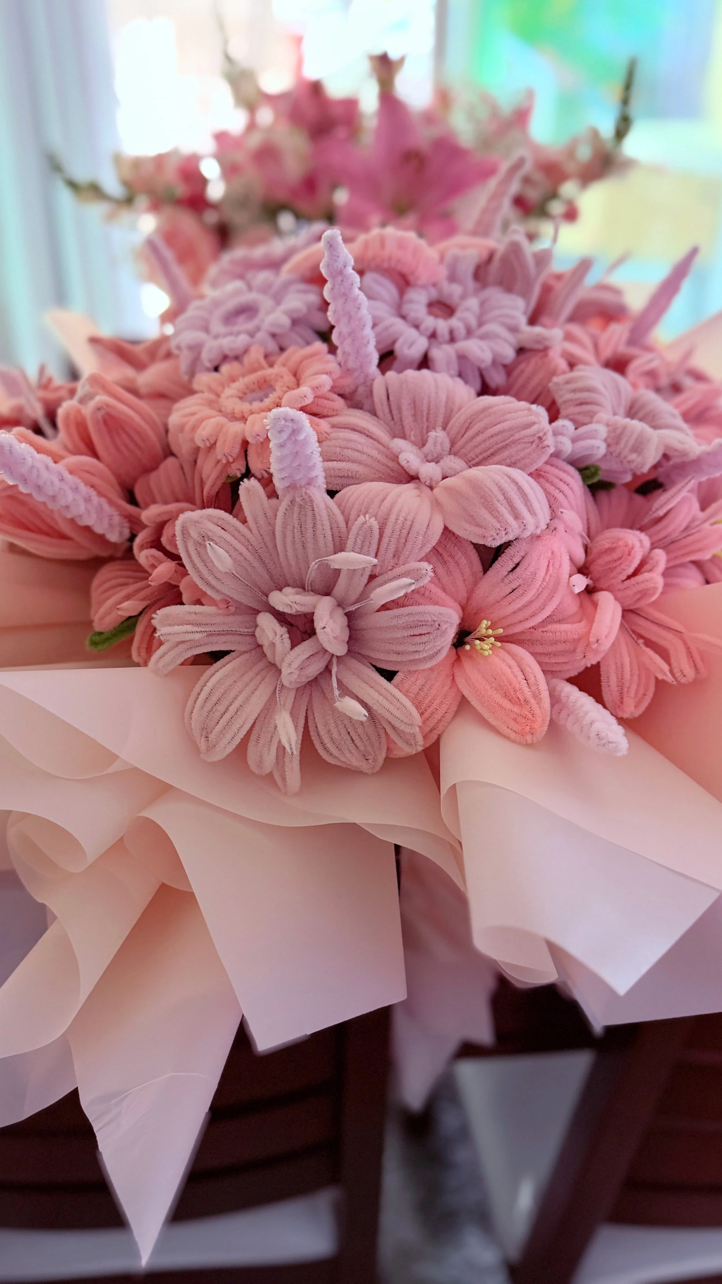 Close-up of a bouquet of pink velvet flowers wrapped in white paper, with a blurred background of flowers and greenery.