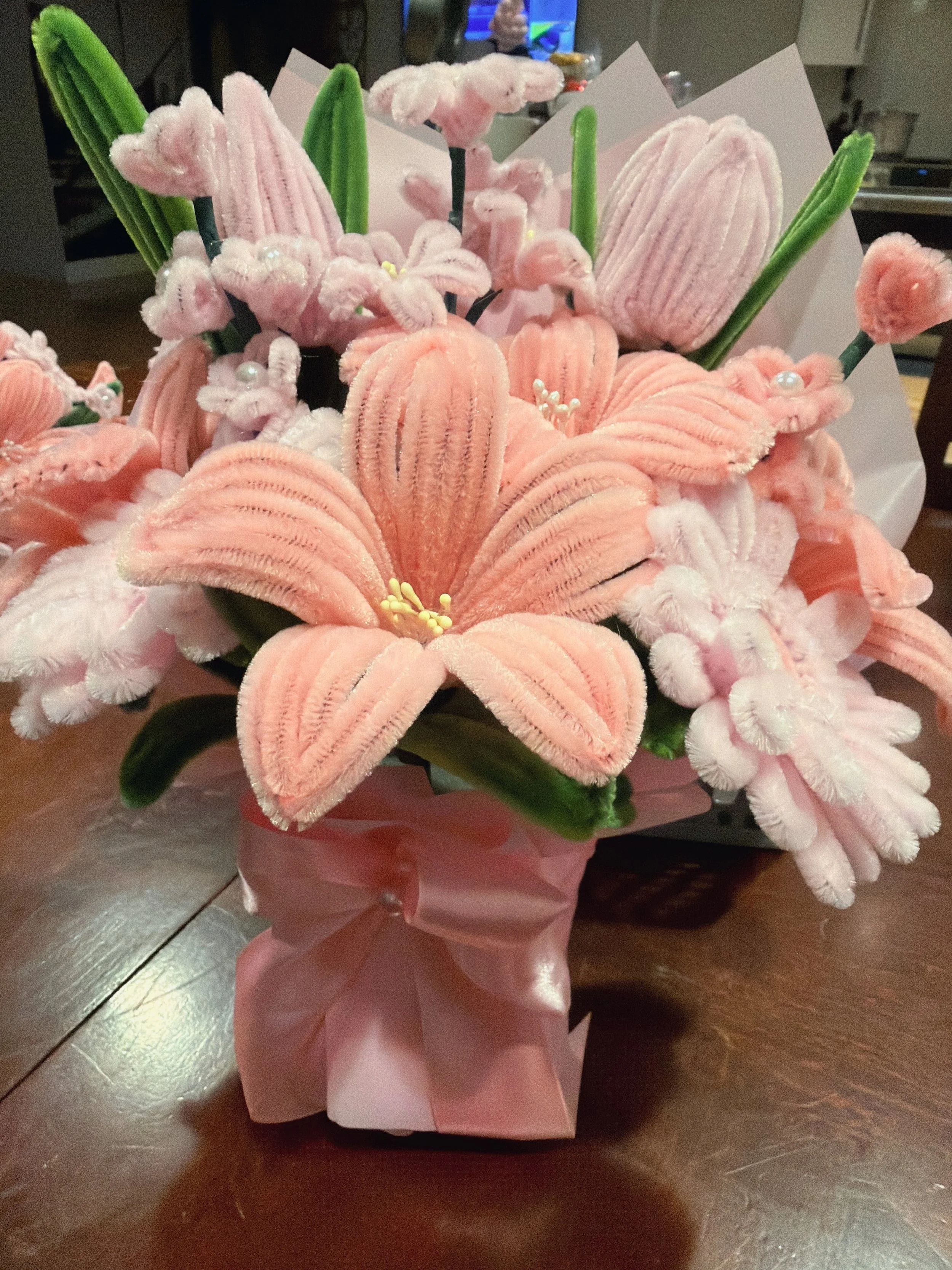 A bouquet of plush toy flowers, including pink lilies, pink roses, and green leaves, arranged together and wrapped in pink paper.