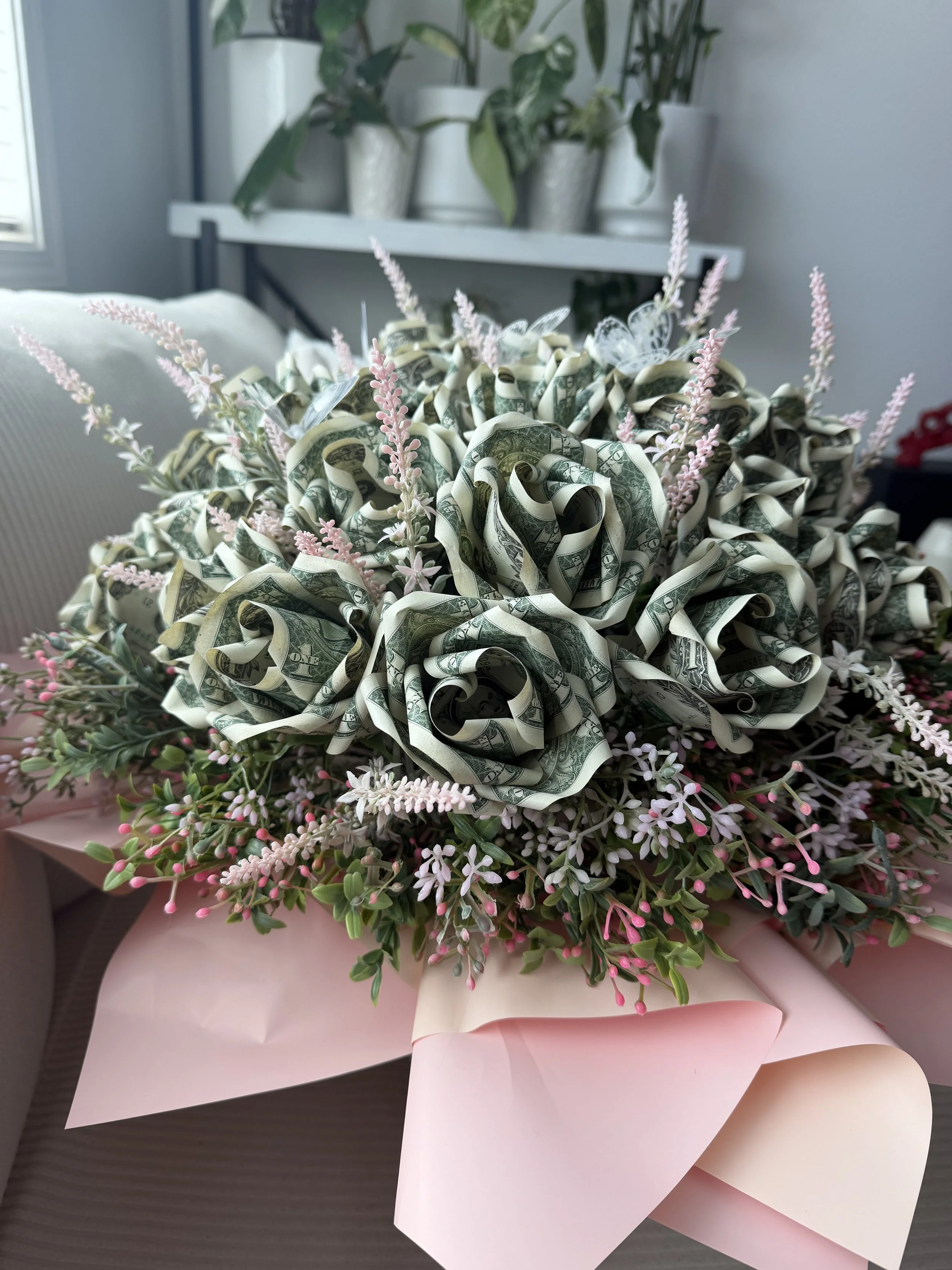Bouquet made of folded U.S. dollar bills with pink and white flowers and green foliage, wrapped in light pink paper.