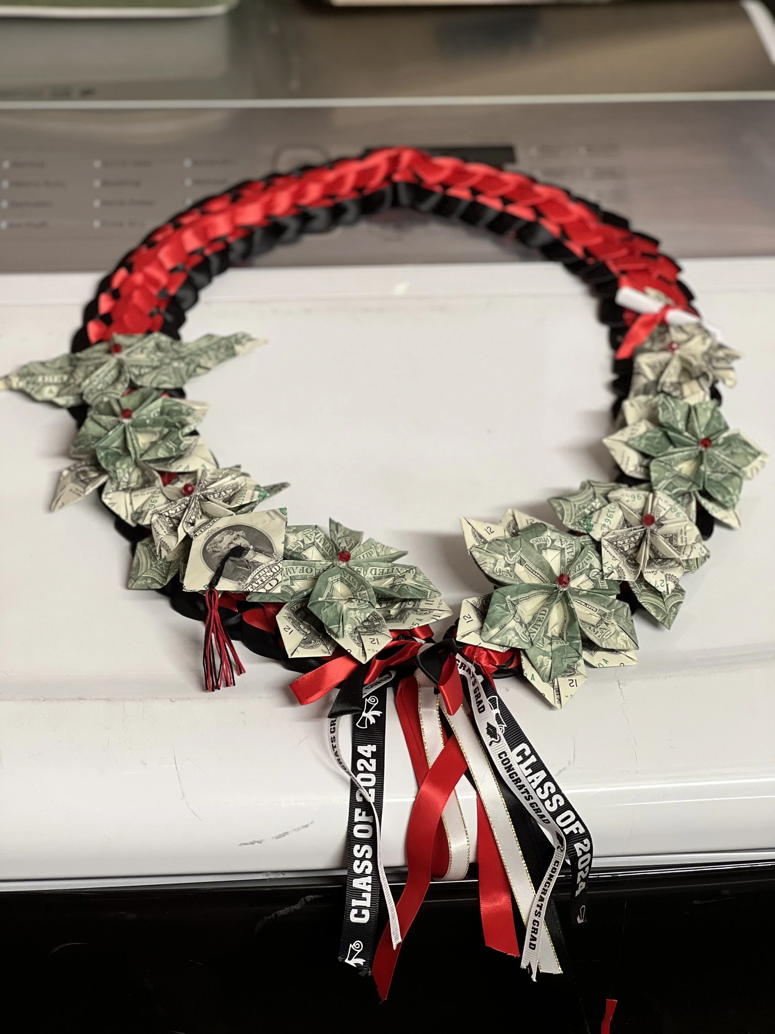 A circular graduation wreath decorated with folded dollar bills, accented with red, black, and white ribbons, and a small tassel.