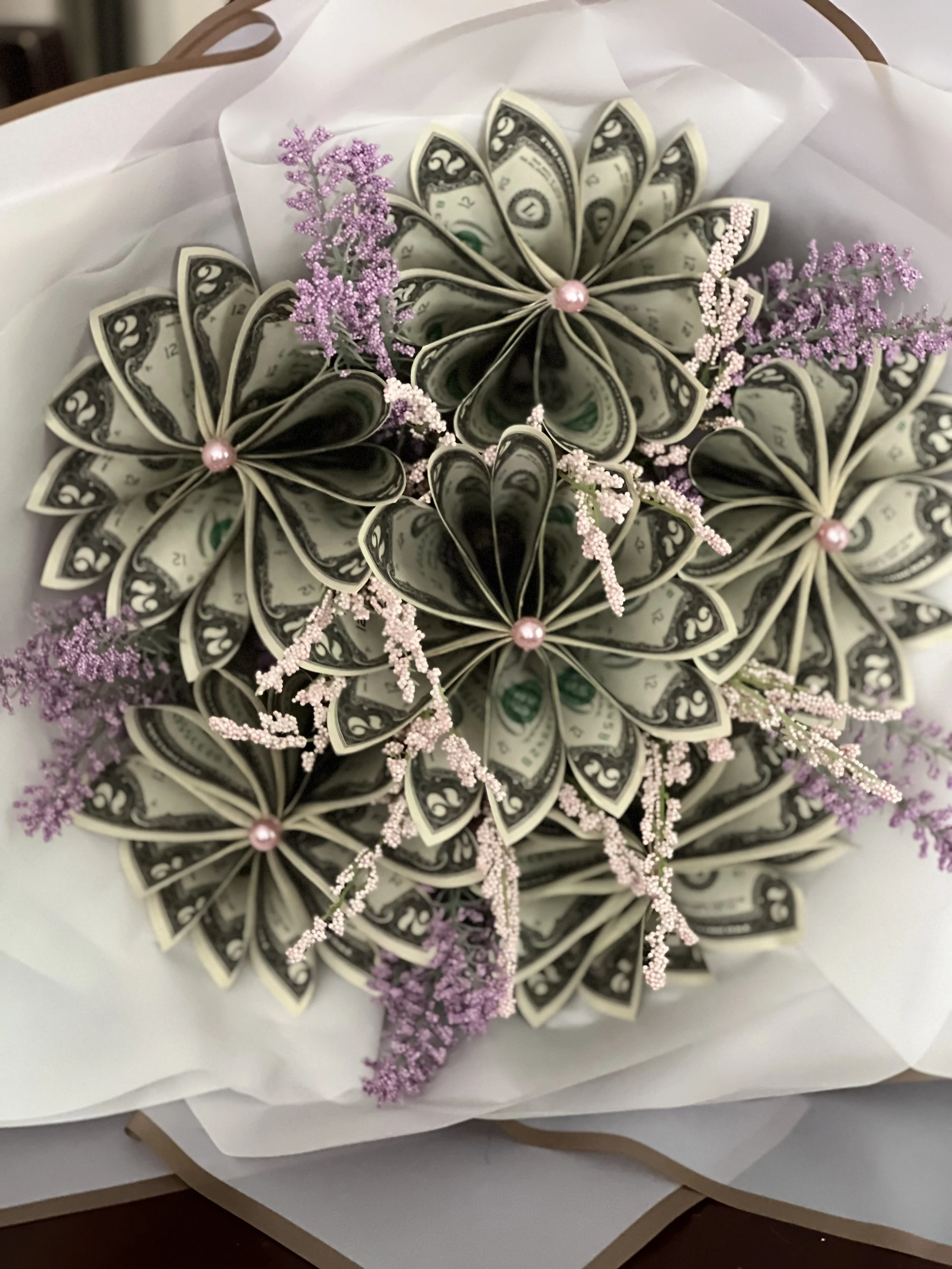 A bouquet made of folded dollar bills resembling flowers, accented with pink and purple artificial small flowers and pink pearl-like beads, wrapped in white paper.