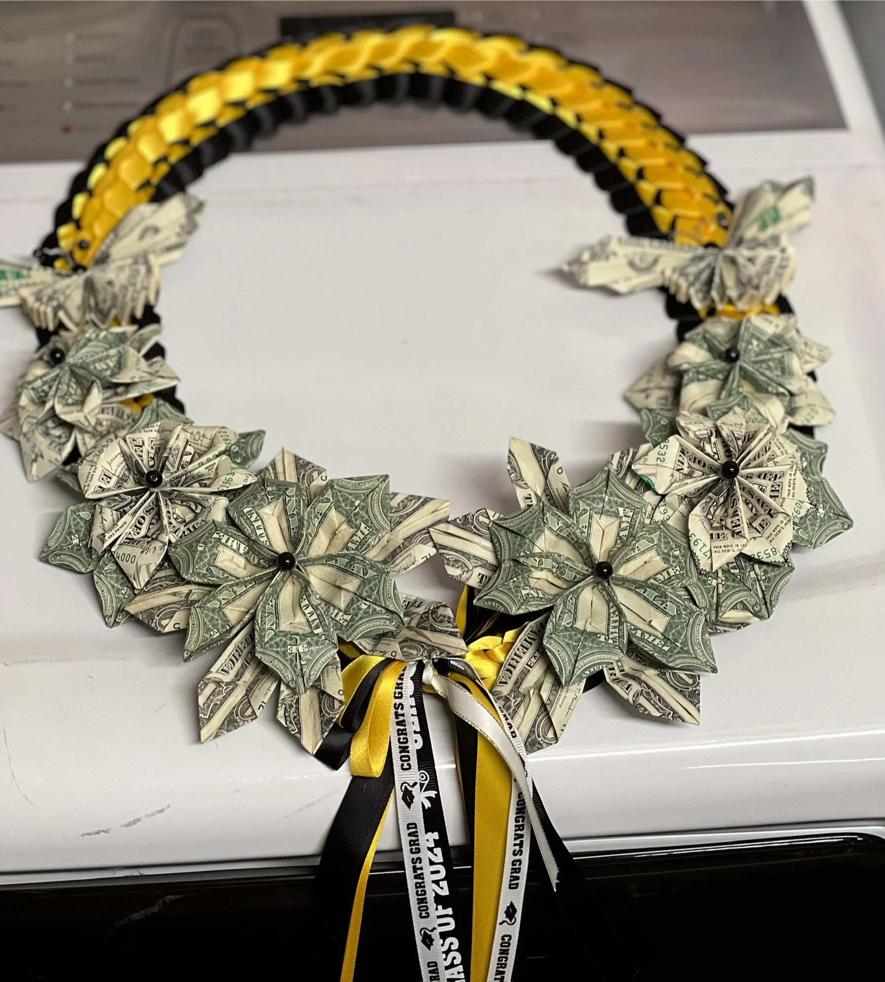 Wreath made of folded dollar bills with black, yellow, and white ribbon accents and small black beads at the centers of the flowers, on a white surface.