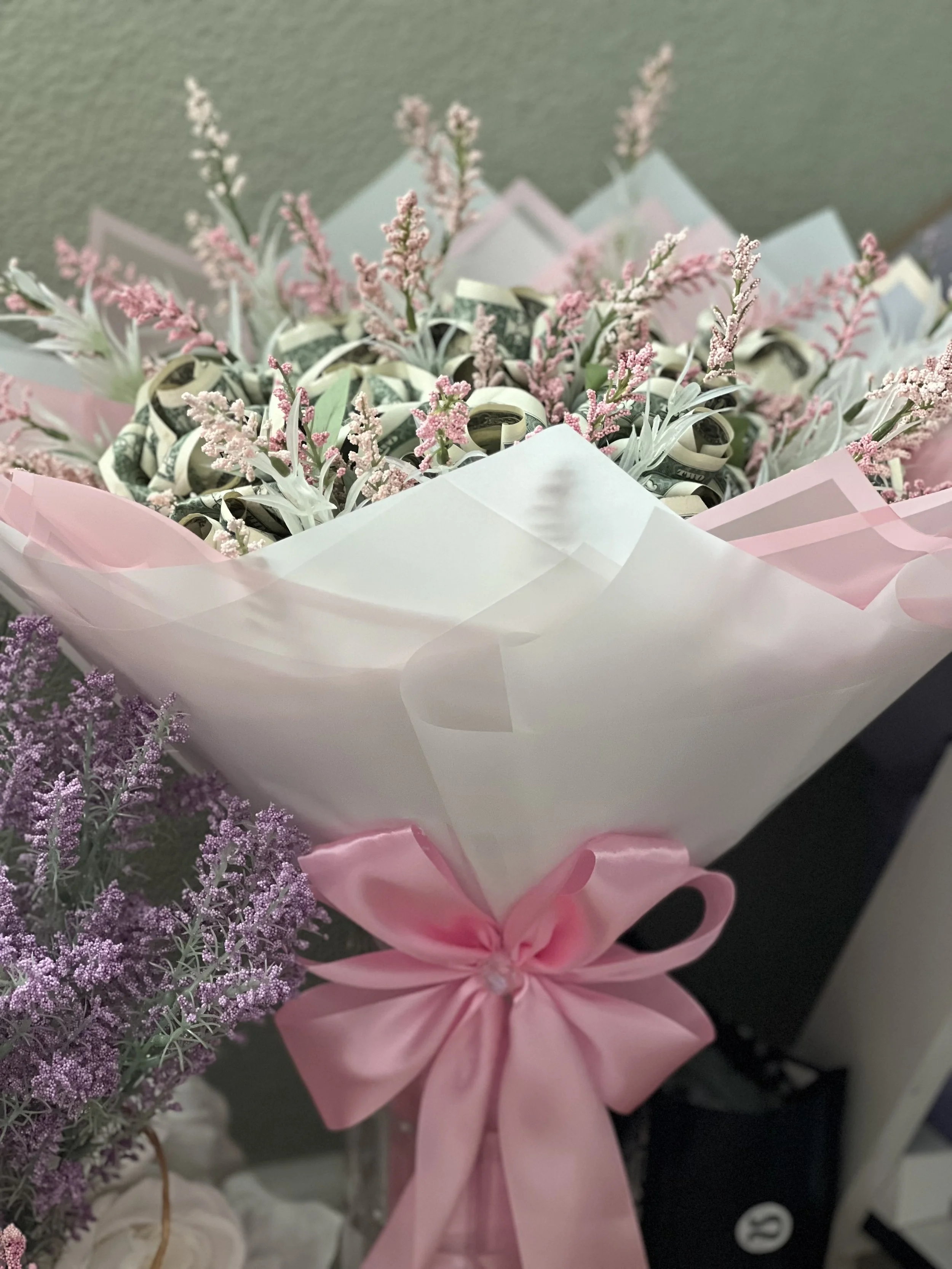 Close-up of a bouquet with pink and purple flowers and money rolled into small bundles, decorated with pink ribbon.