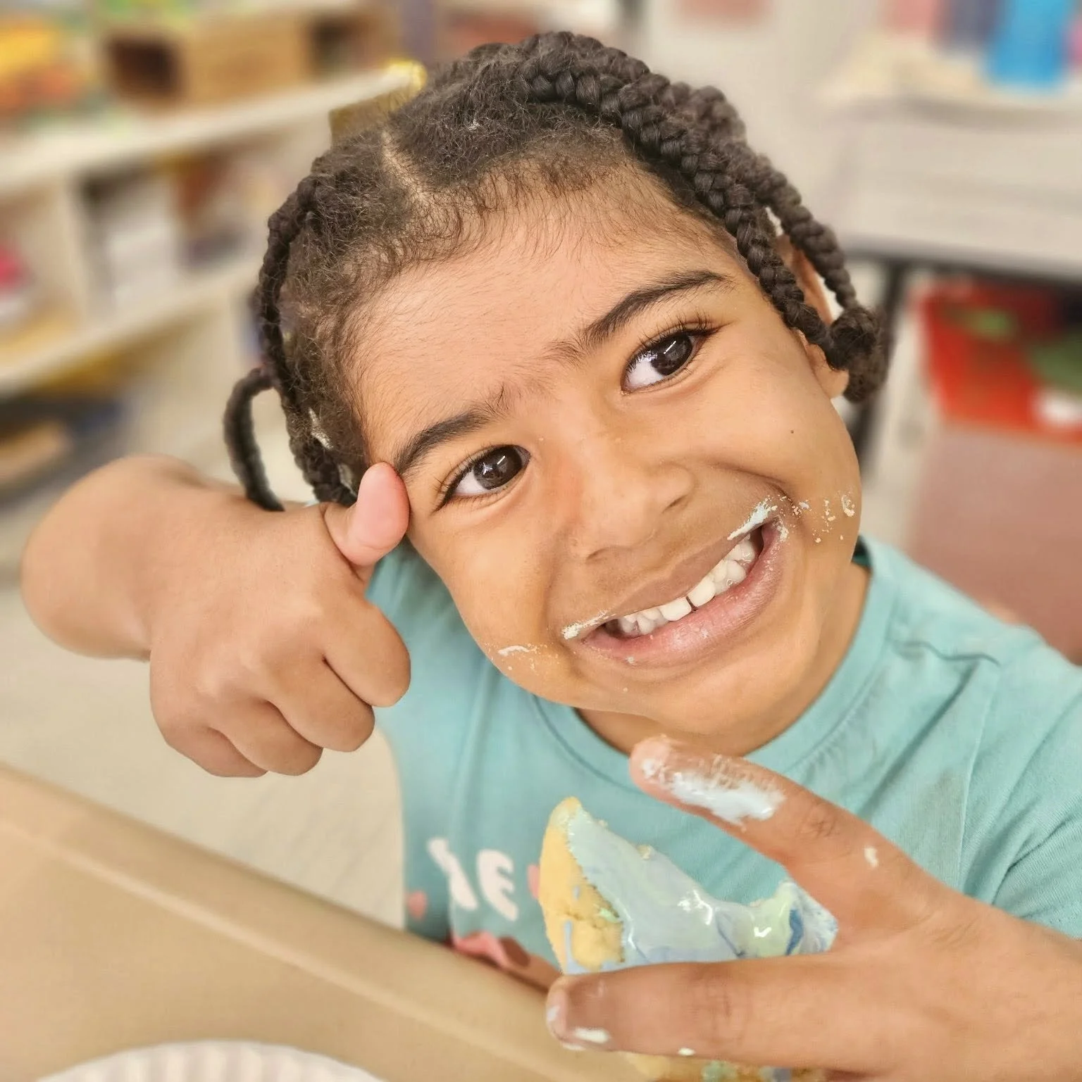 Young girl with braided hair smiling and giving a thumbs up, with frosting on her face and hands, and a piece of cake in front of her. WHCCNS