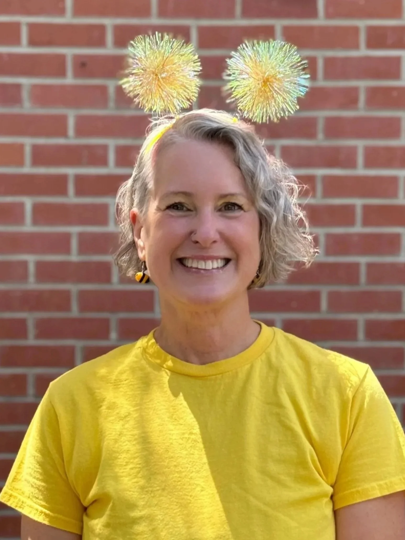 A woman with short, curly gray hair smiling and wearing a bright yellow shirt, standing in front of a red brick wall with two yellow and black bee headbands on her head.