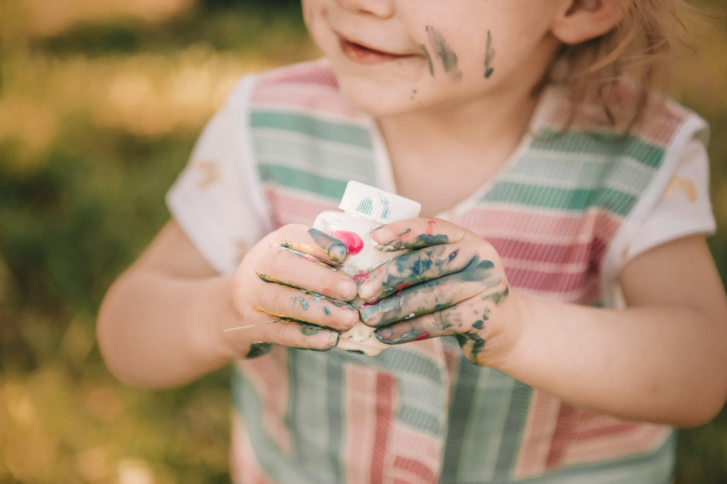 Close-up of a young child with painted fingers holding a small container of paint, with paint on their face, wearing a colorful striped shirt, outdoors with blurred background.