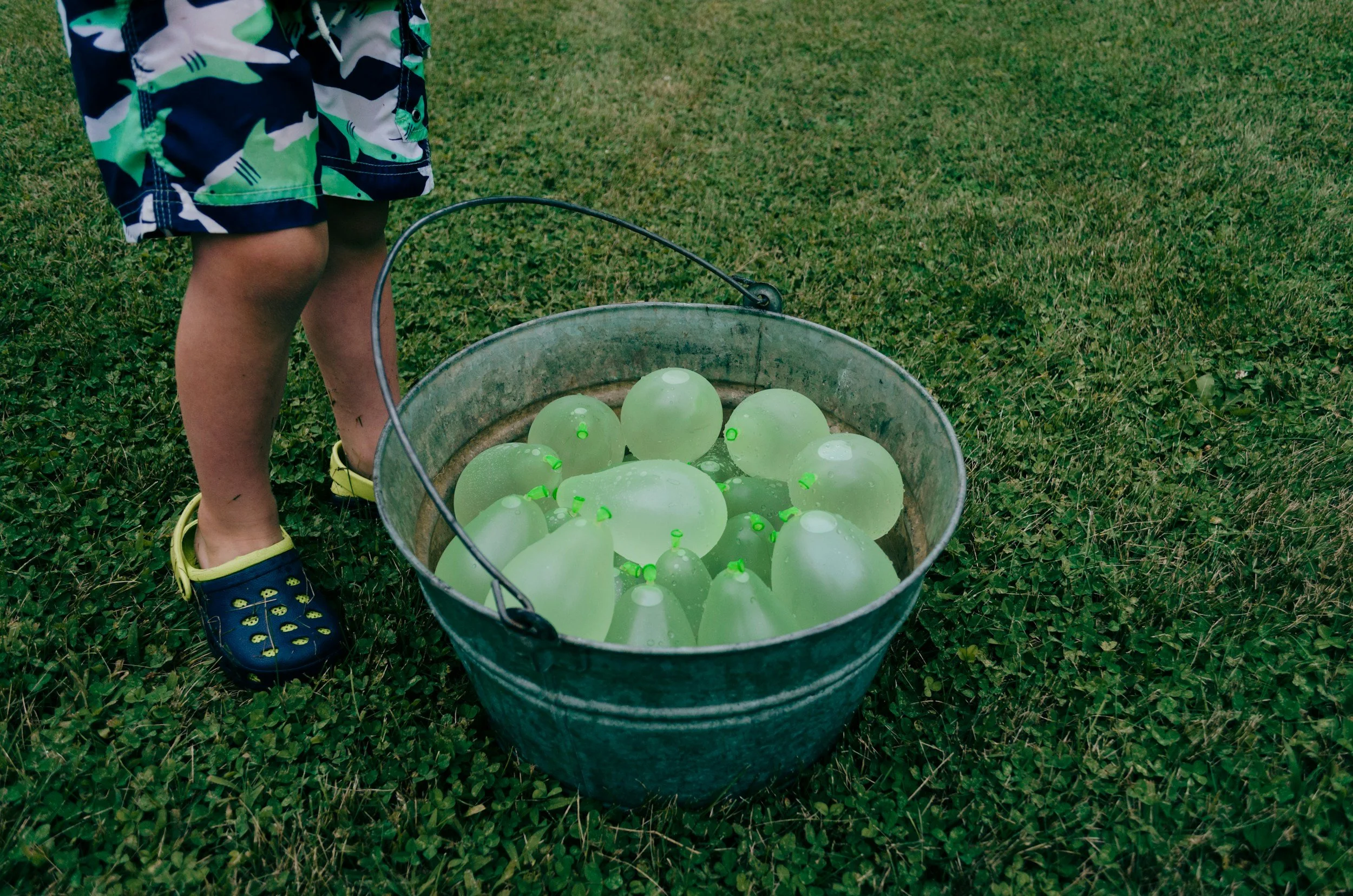 Child standing on grass wearing blue shoes with yellow smiley faces, near a metal bucket filled with water and greenish-white balloons.
