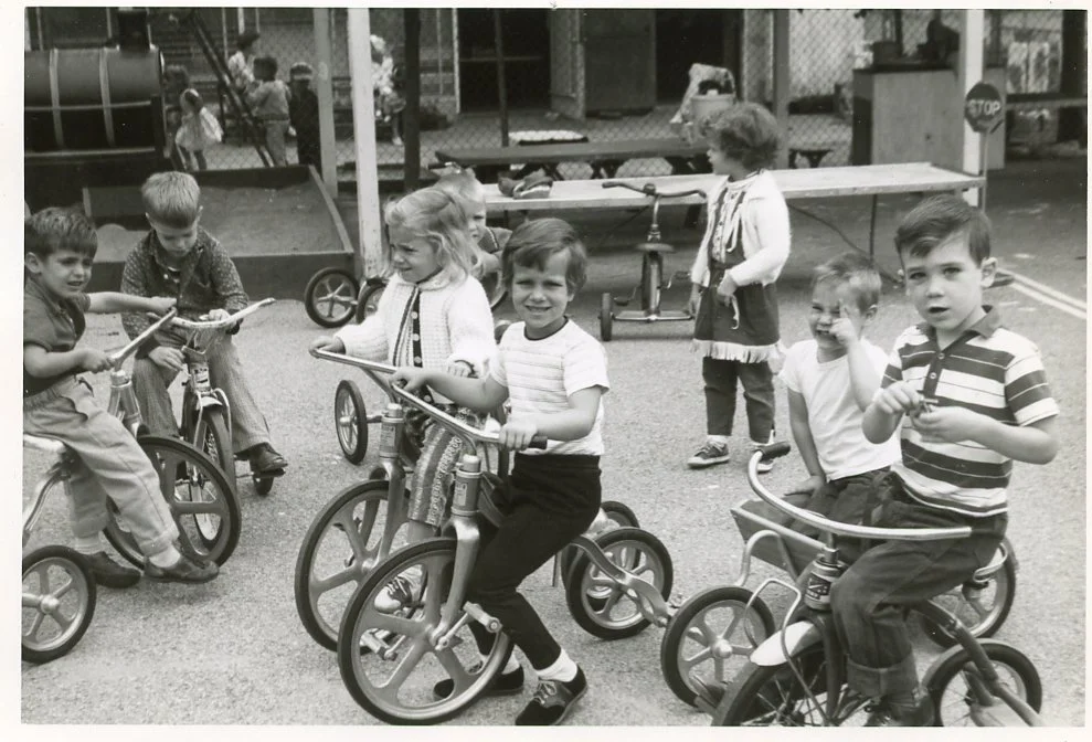 Young children riding tricycles and bicycles in a playground, with a fence and play area in the background at WHCCNS
