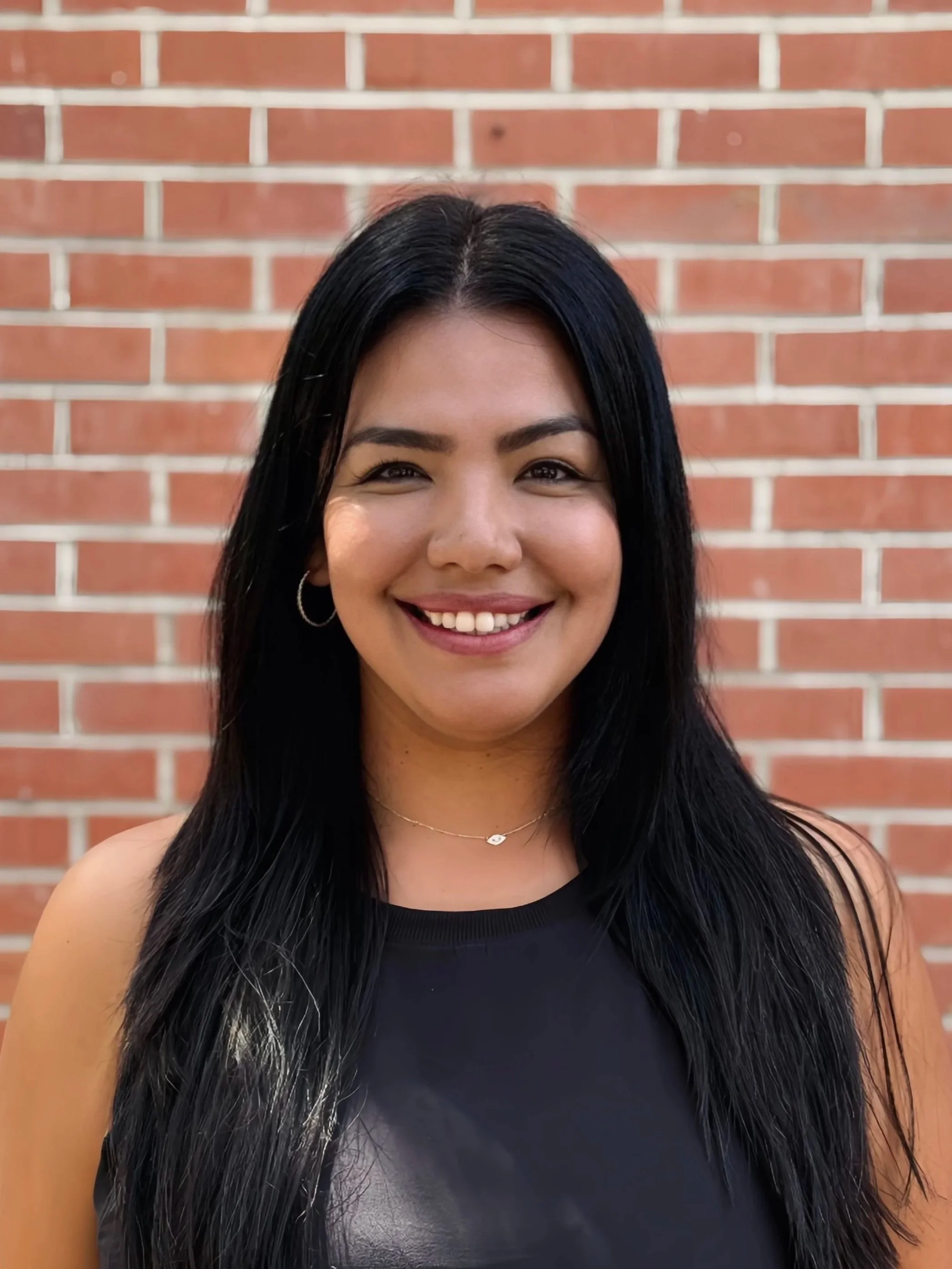A woman with long black hair, wearing hoop earrings, a delicate necklace, and a black top, smiling in front of a brick wall background.