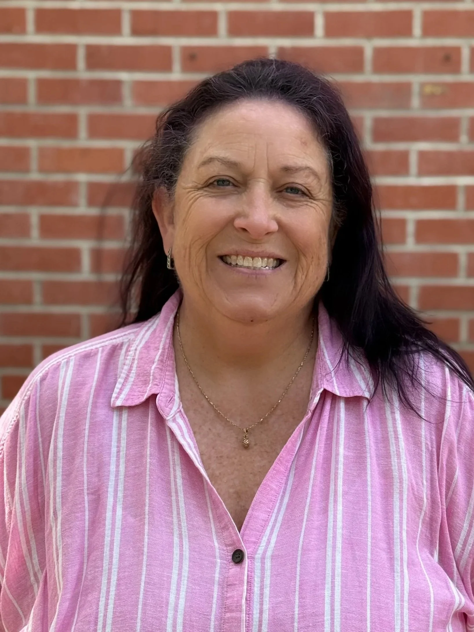 A middle-aged woman with dark brown hair, wearing a pink striped shirt, standing in front of a red brick wall, smiling.