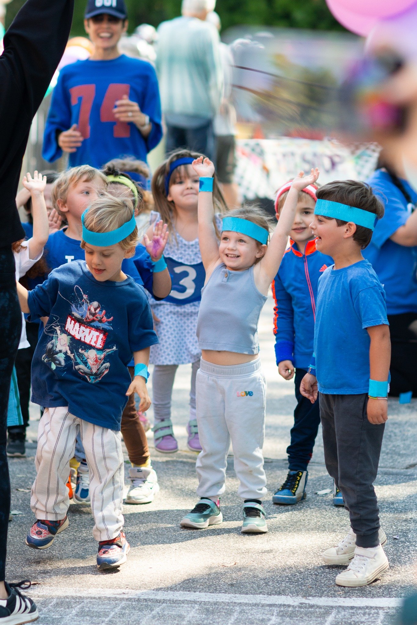 Group of children at a fun outdoor event, some wearing blue headbands, smiling and raising their hands, with a woman in a blue shirt and a woman with white hair in the background. Jog -a-thon at WHCCNS