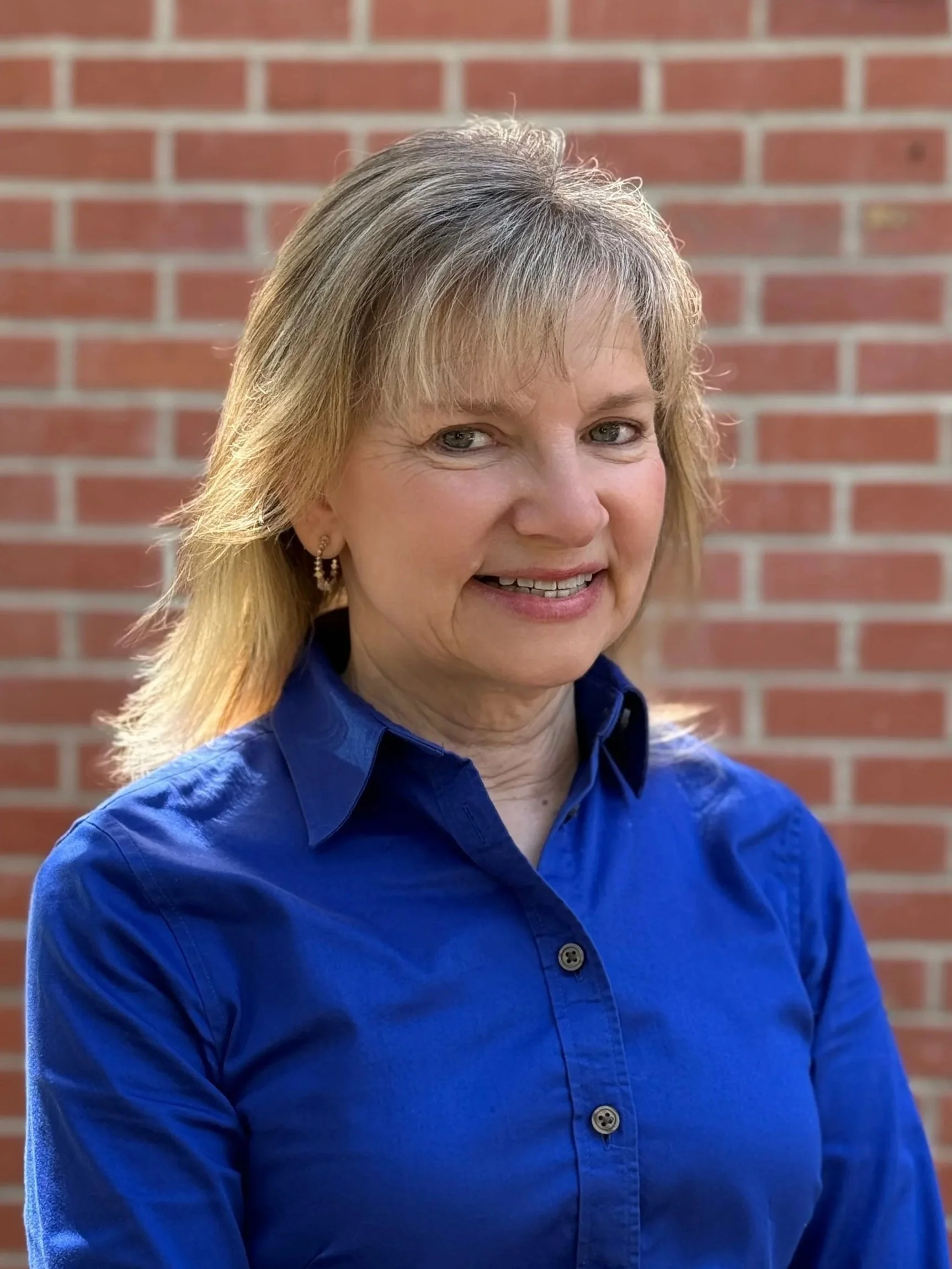 A woman with blonde hair wearing a blue shirt, smiling, standing outdoors against a red brick wall.